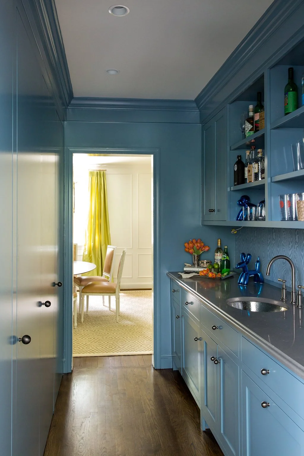 A narrow kitchen with blue cabinets and open shelves, a stainless steel countertop and sink, and decorative items like balloons and flowers. Sunlight comes from a room with a table, chairs, and yellow curtains.