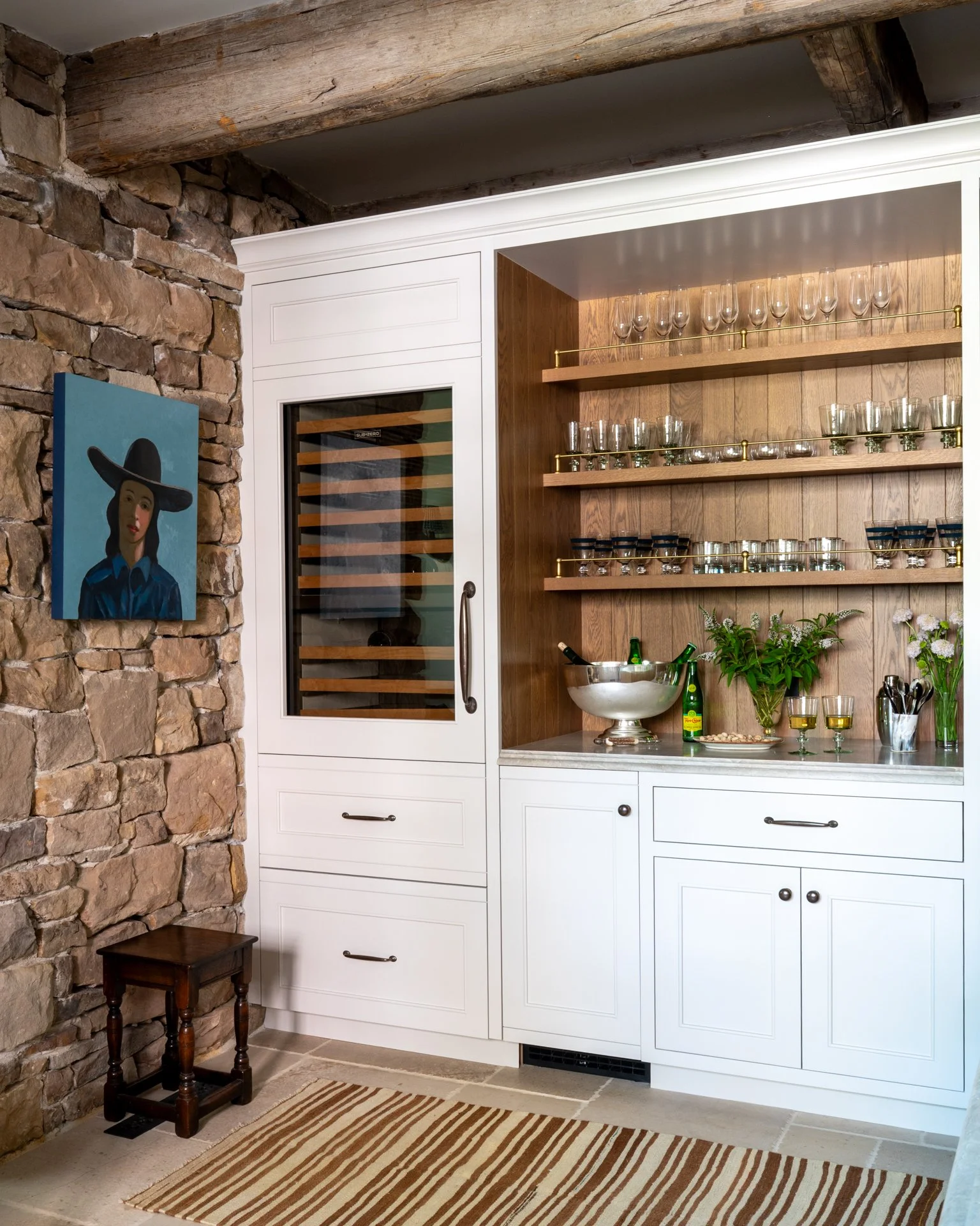 A cozy kitchen corner with a rustic stone wall, a wooden beam overhead, and white cabinetry. There are glassware on the shelves, a metal bowl with green bottles, and flowers on the countertop. A portrait of a woman in a large hat is hung on the stone