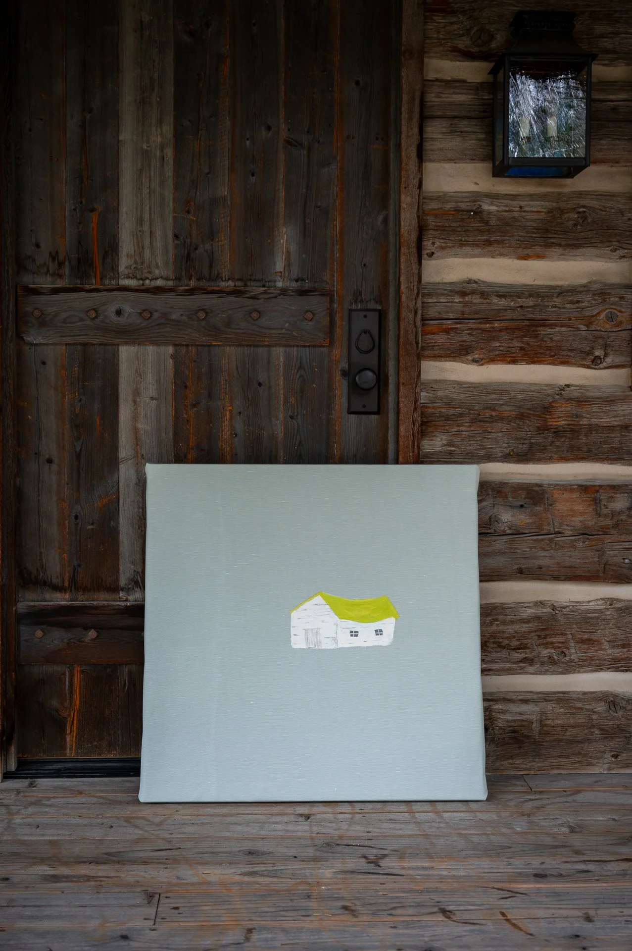 A canvas with a painted white house with a green roof, placed on a wooden porch against a rustic wooden door and logs wall.