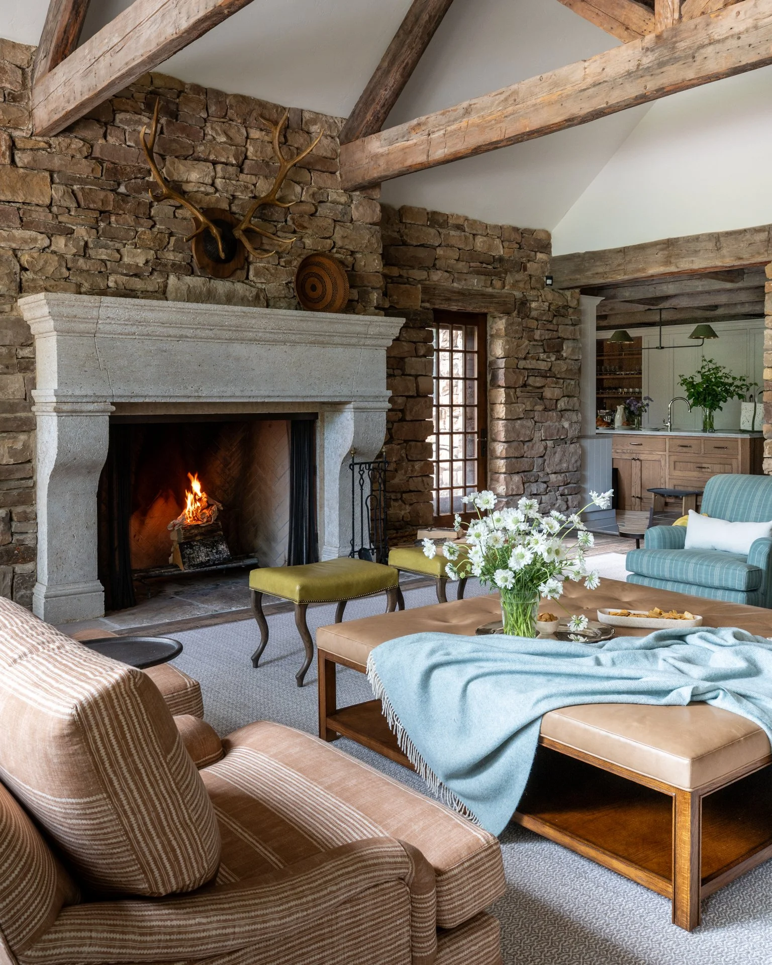 Living room with stone fireplace, wooden beams, and furniture including a striped armchair and a sofa with a throw blanket, a glass vase with white flowers on a wooden table, and a doorway leading to a kitchen area with plants and hanging lights.