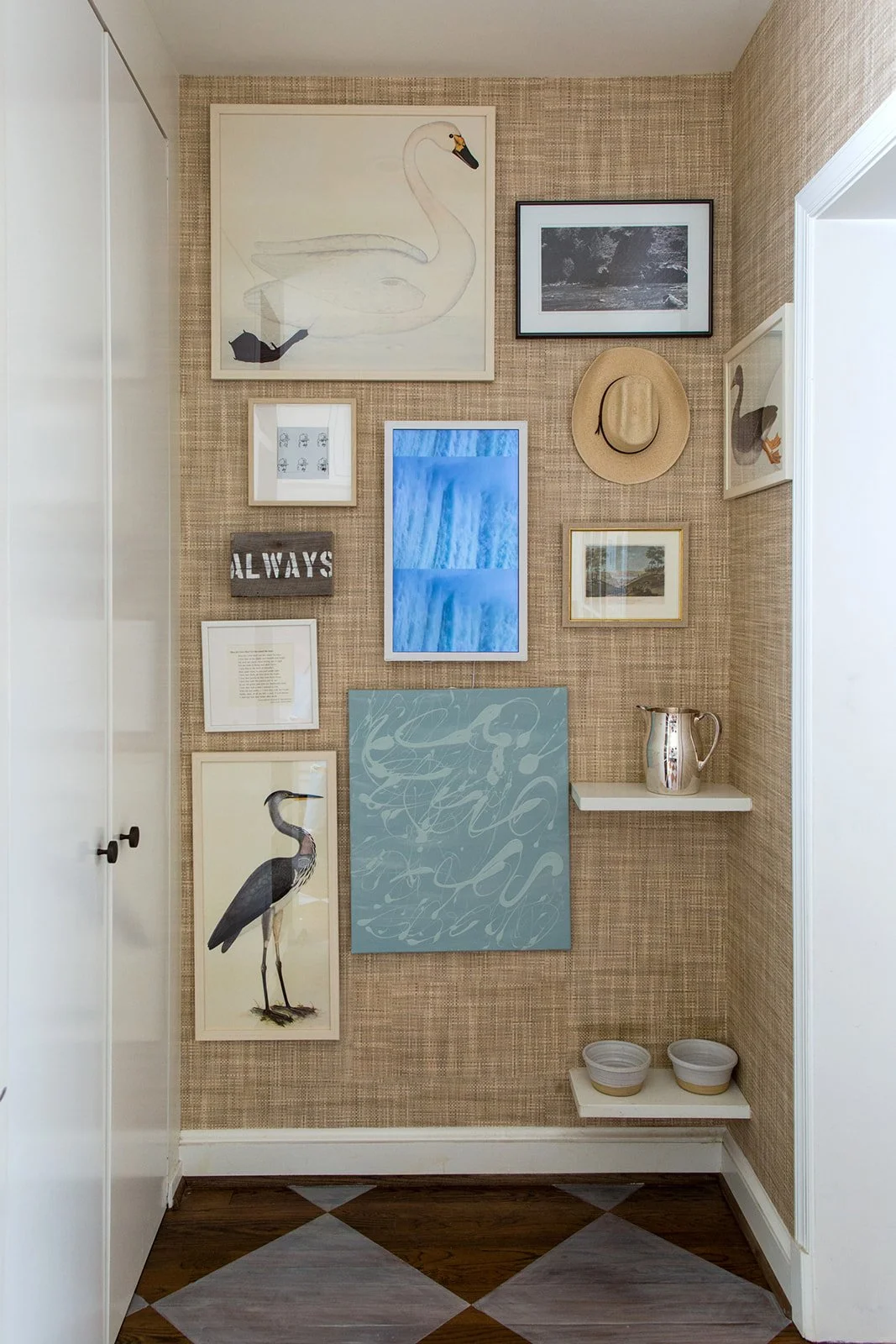 Wall decorated with various framed artworks, a straw hat, a metal pitcher on a white shelf, and two small bowls on a lower shelf, with a partial view of a white door and wood floor.