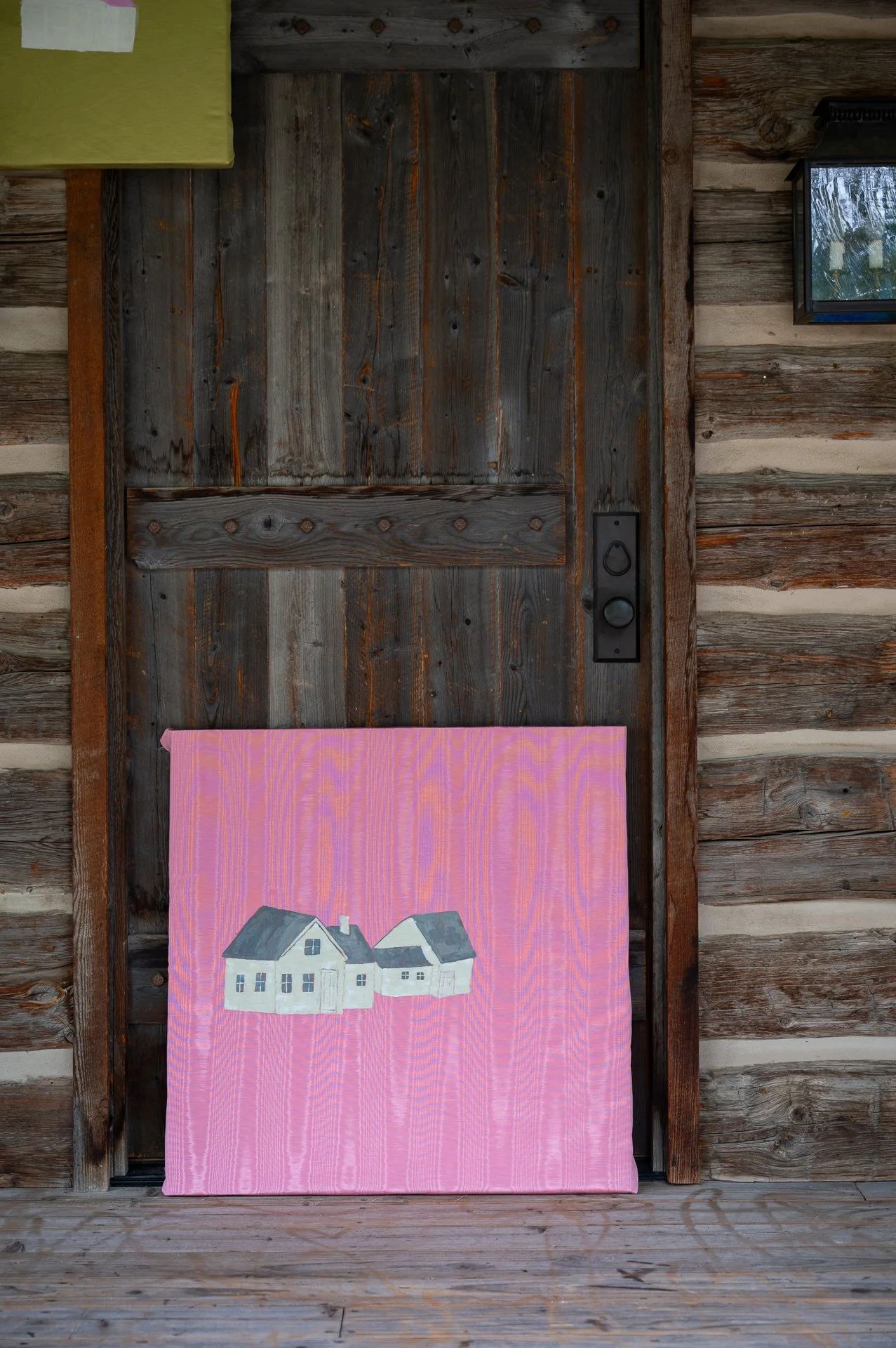 A painting of two houses on a pink background, leaning against a wooden wall outside a rustic cabin.