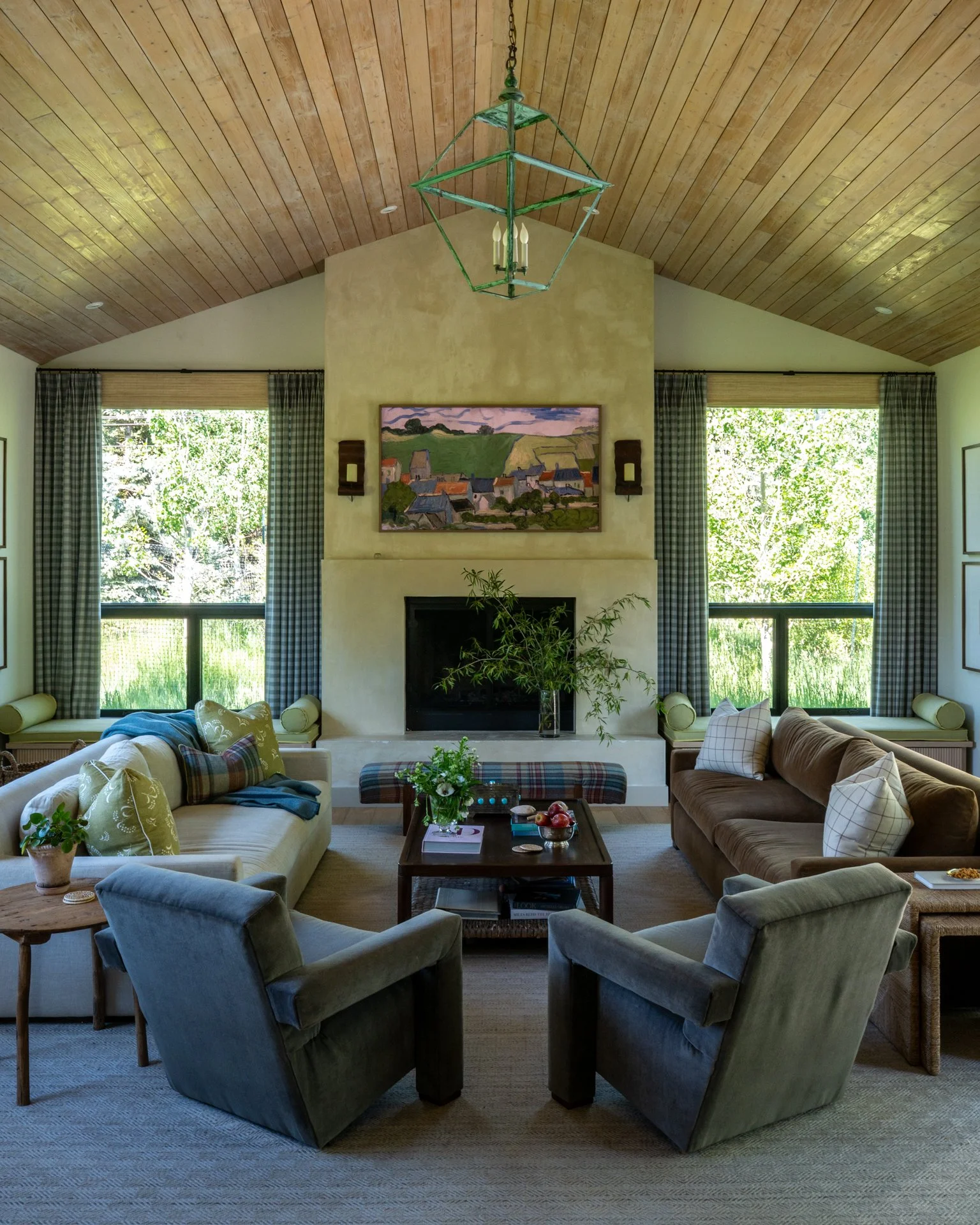 A spacious living room with a high wooden vaulted ceiling, a fireplace flanked by two large windows with blue checkered curtains, and comfortable sofas and chairs arranged around a central coffee table, decorated with plants, books, and snacks.