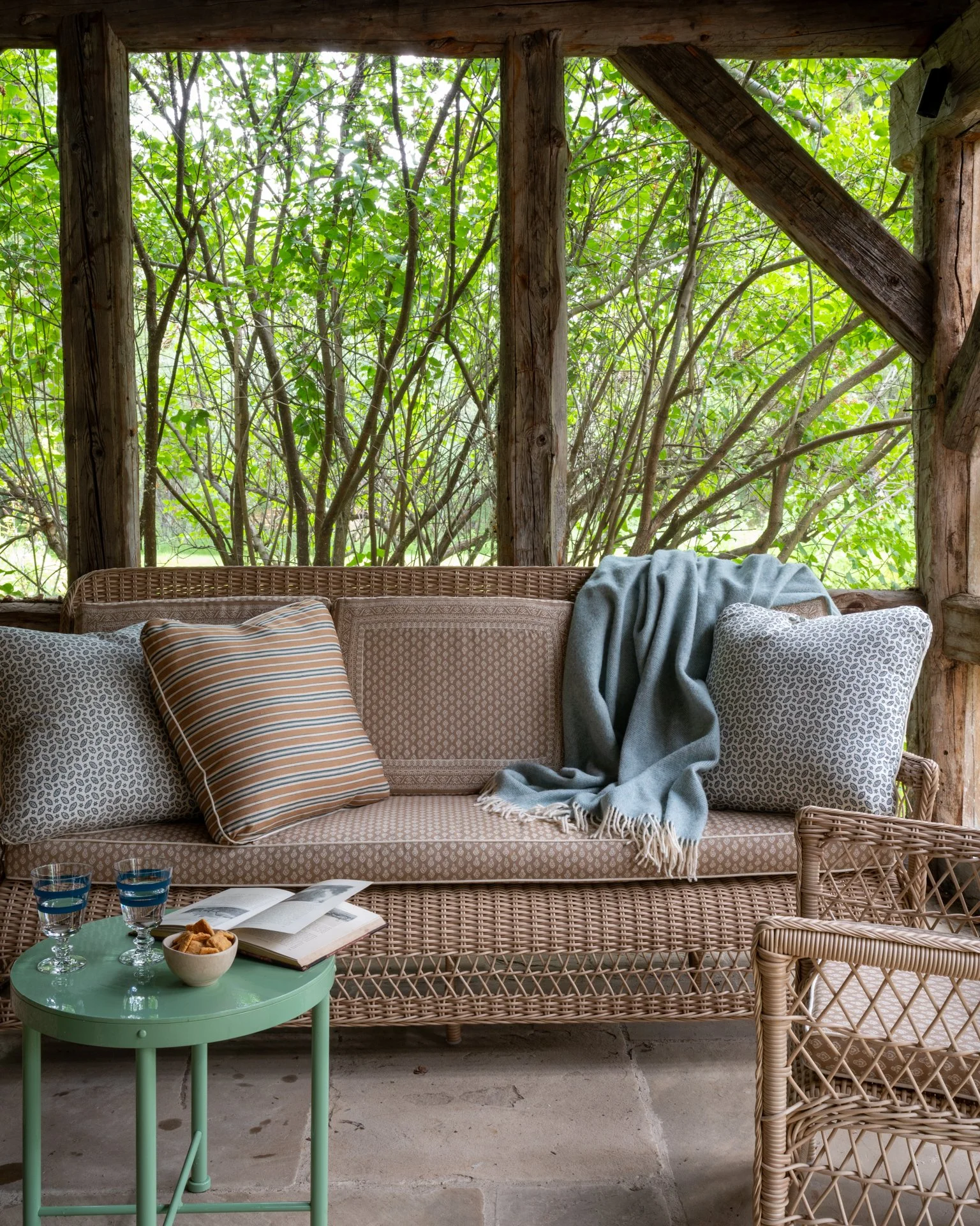 Porch seating area with wicker sofa with patterned pillows and blanket, surrounded by greenery visible through the openings in the porch wall, with a green side table holding glasses and snacks.
