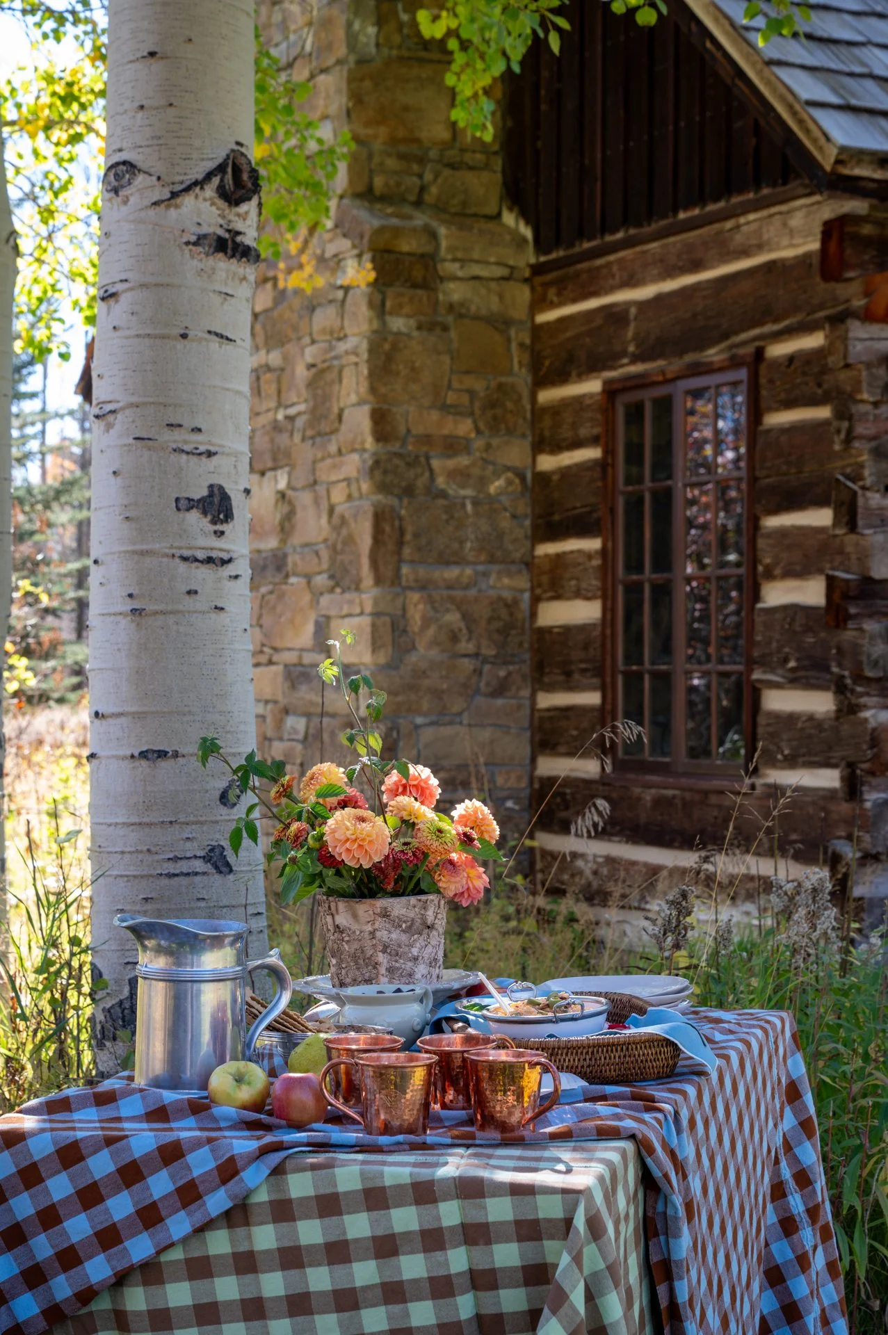 Outdoor table set with a checkered tablecloth, flowers, apples, copper mugs, and dishes in front of a rustic log cabin with a stone chimney and window, surrounded by trees and greenery.