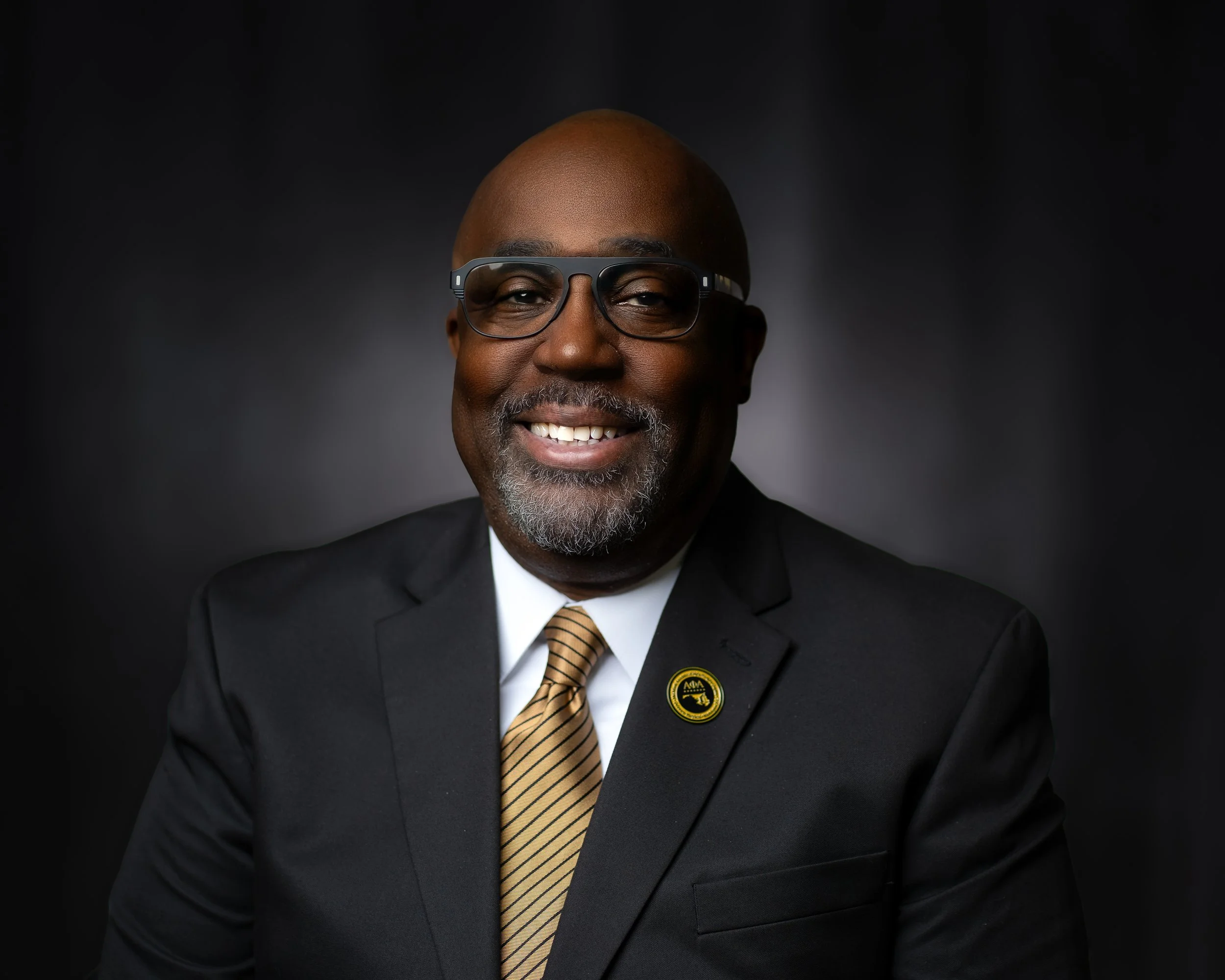 Portrait of a smiling African American man wearing glasses, a black suit, white shirt, and yellow striped tie, against a dark background.