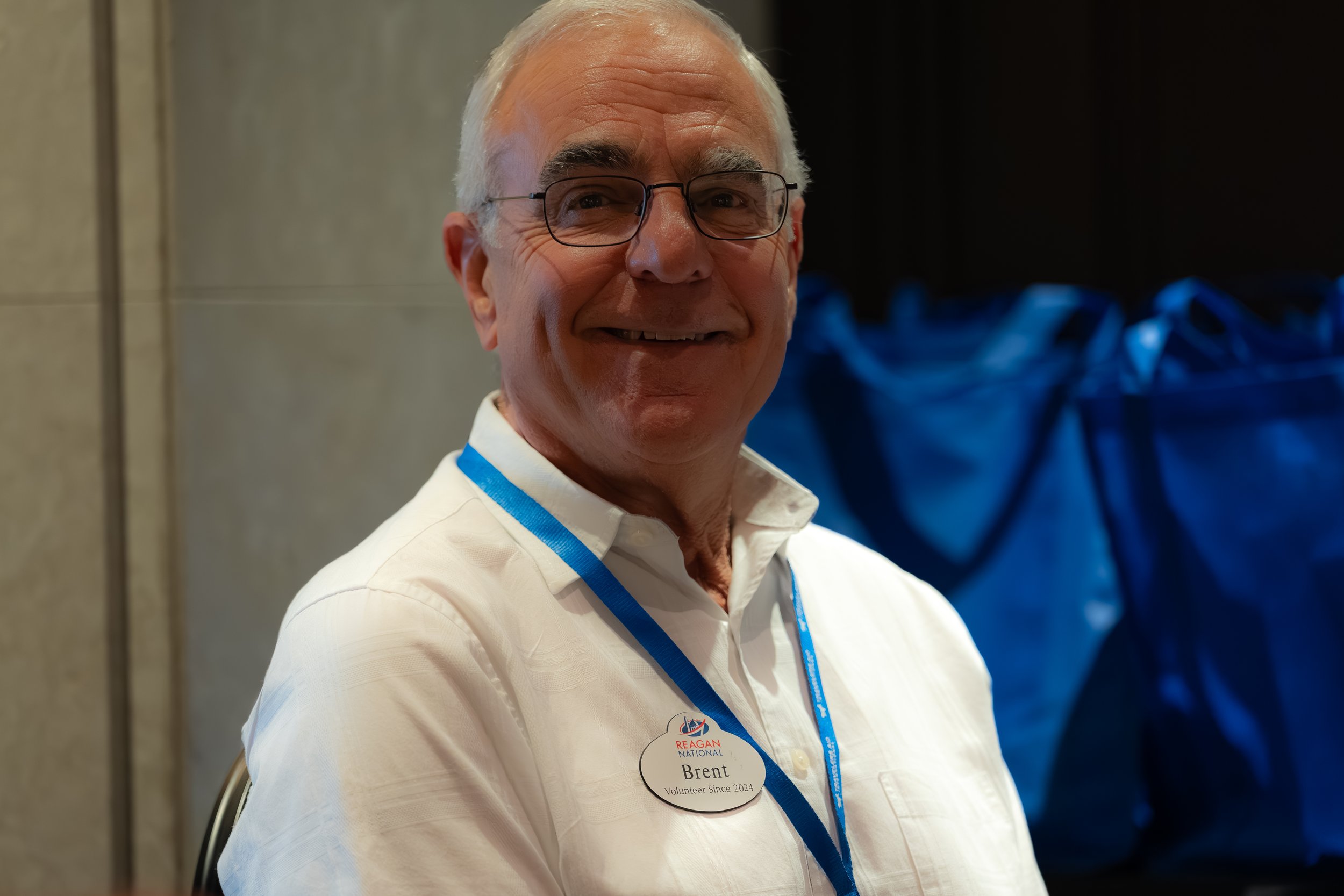 Smiling elderly man wearing glasses and a name tag, sitting indoors.