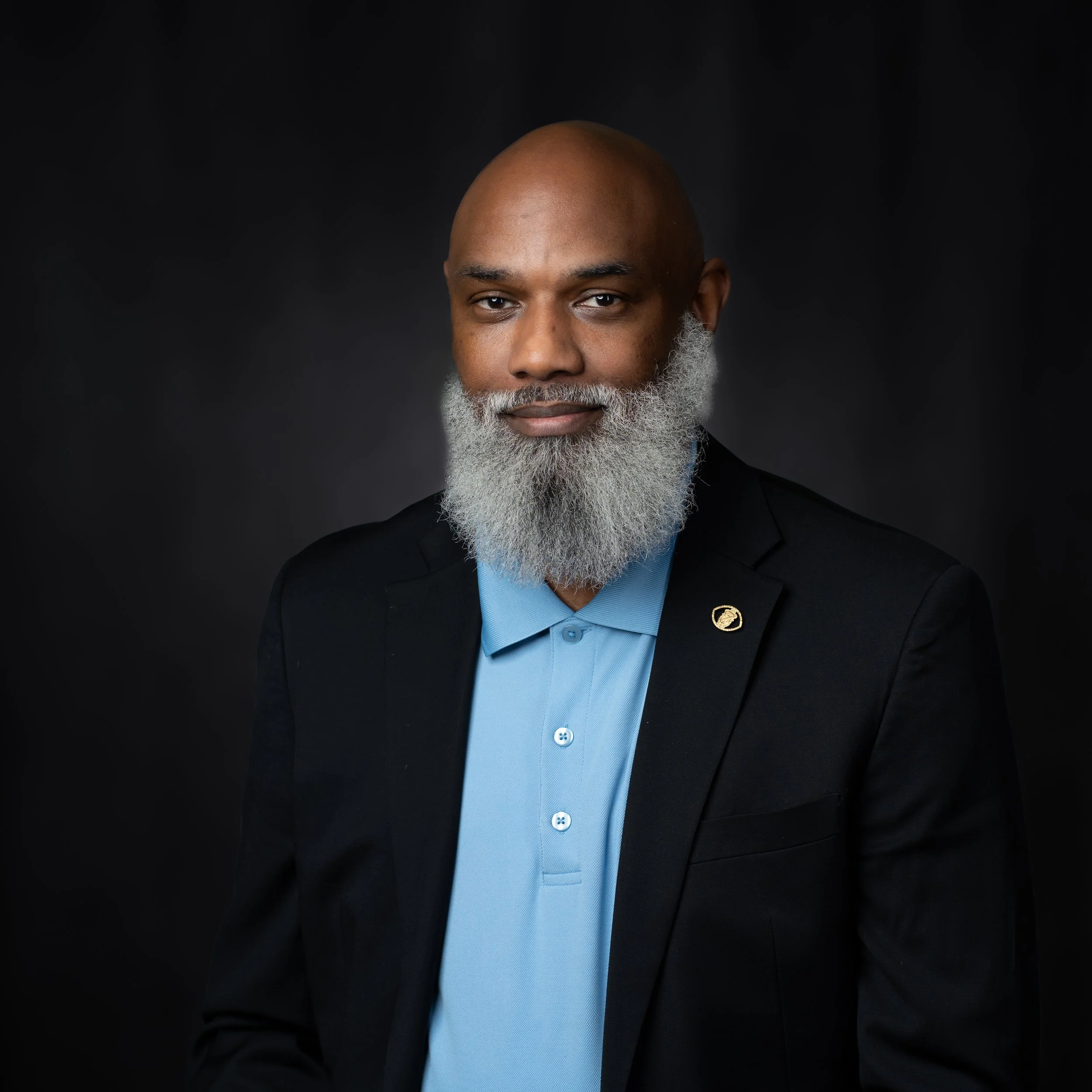 A middle-aged man with a bald head and a long, gray beard, wearing a navy blazer with a pin and a light blue collared shirt, posing against a black background.