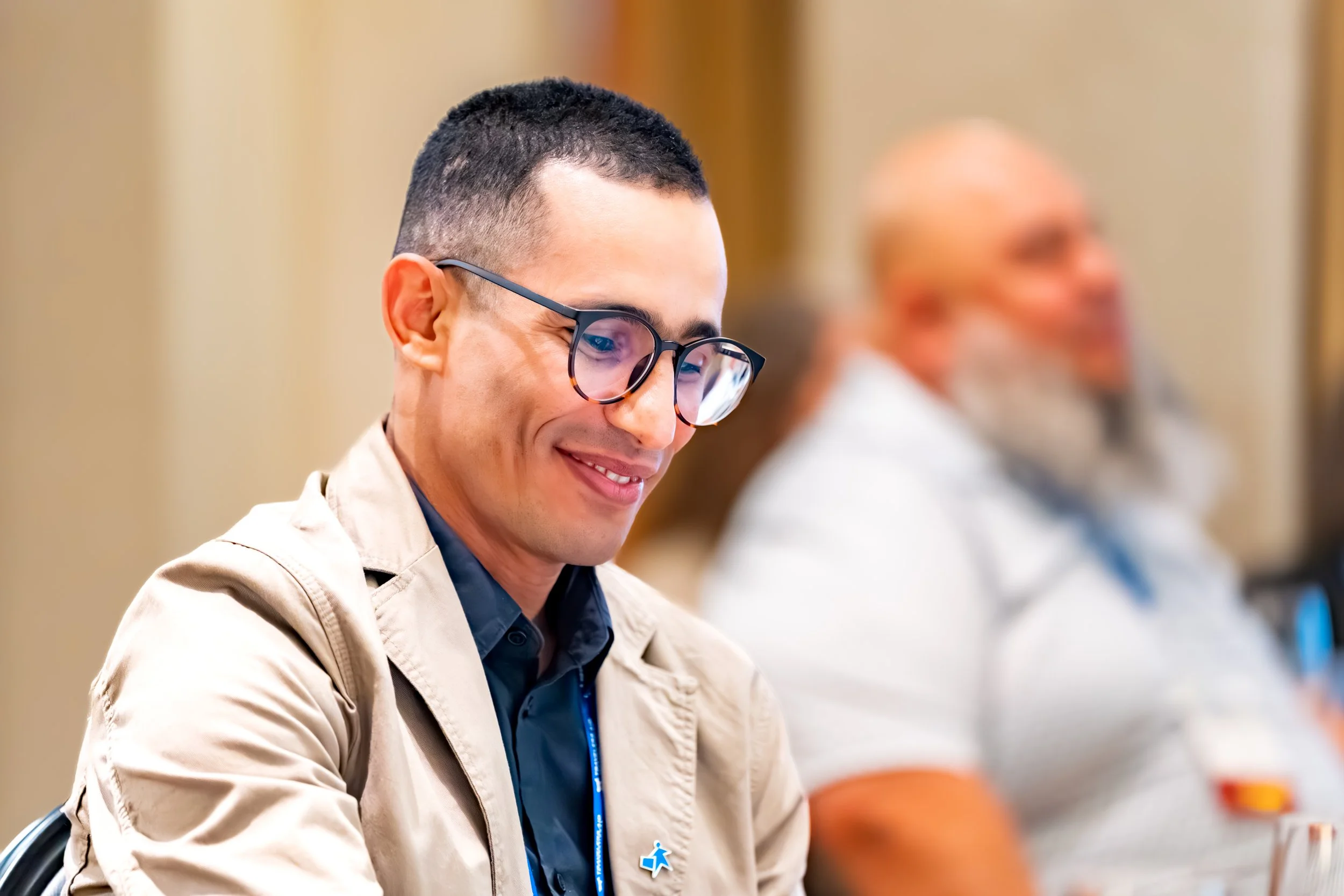 A young man with short dark hair and glasses sitting in a conference, smiling while looking down, with an older man with glasses and a beard blurred in the background.