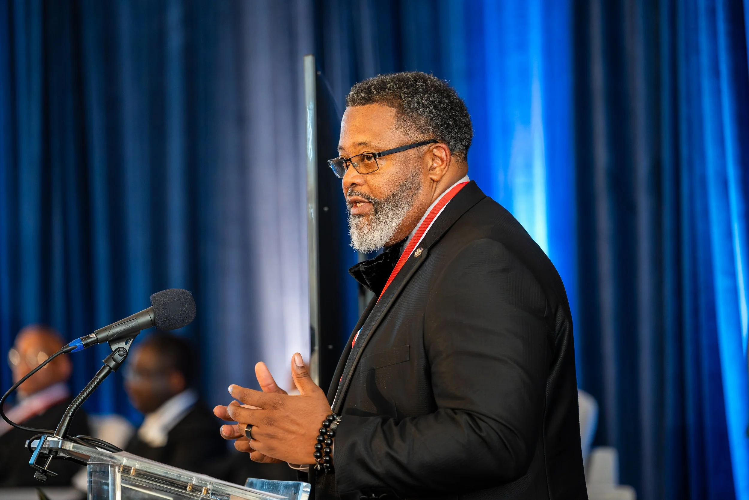 A man in a black suit and glasses speaking at a podium with a microphone in front of a blue curtain, with other formally dressed people seated in the background.