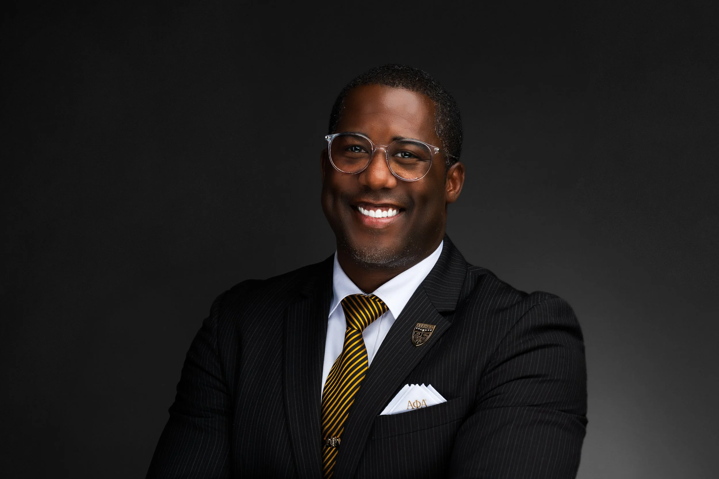 Professional portrait of a smiling African American man in a business suit, glasses, with a black background.