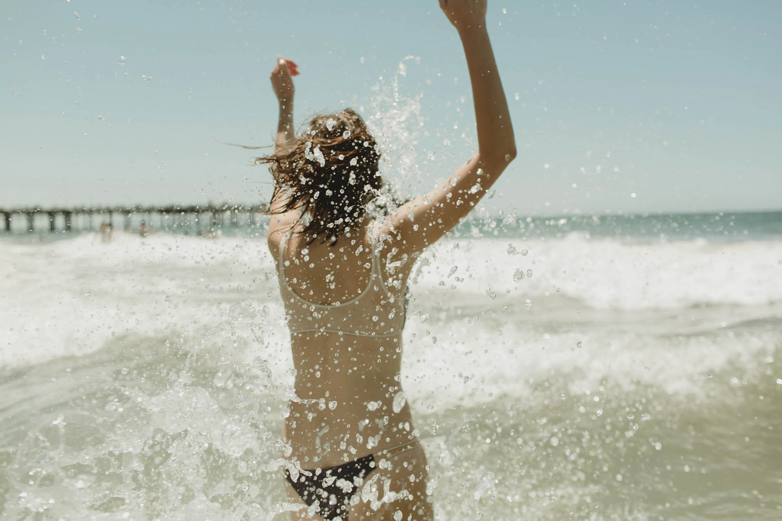Woman in a black bikini playing in the ocean waves near a pier on a sunny day.