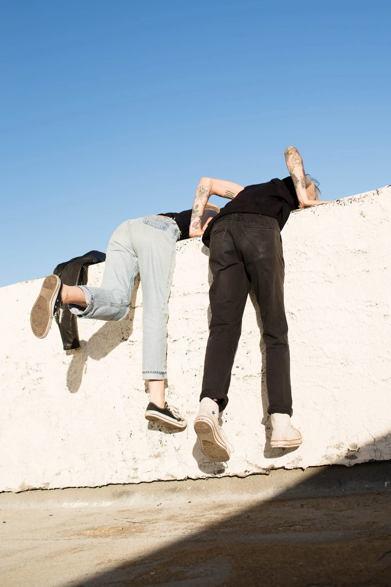 Two people hanging onto the top of a white wall, with one person climbing up and another leaning over.