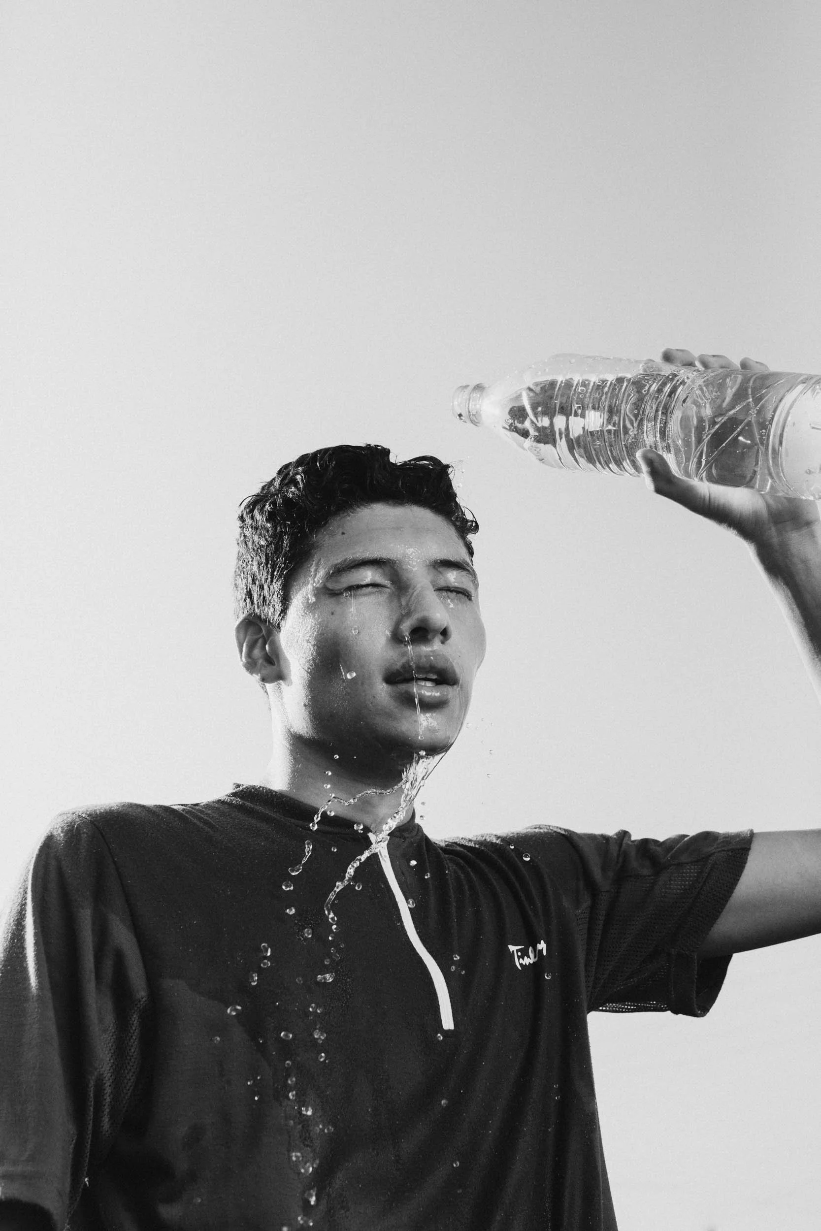 A young man in athletic clothing is pouring water over his face from a plastic water bottle, with water dripping down his face.