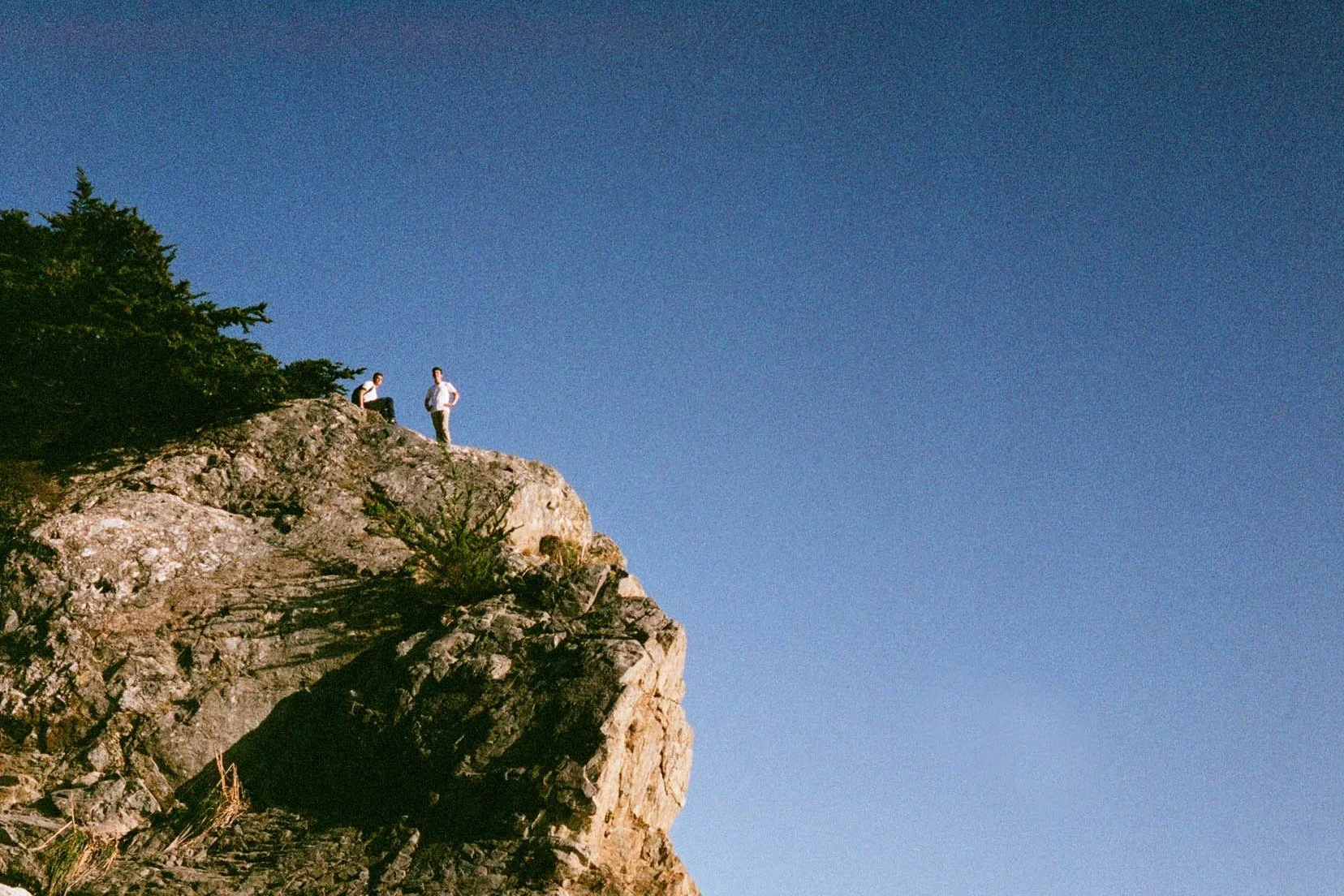 Two people standing on a rocky cliff edge with a clear blue sky in the background.