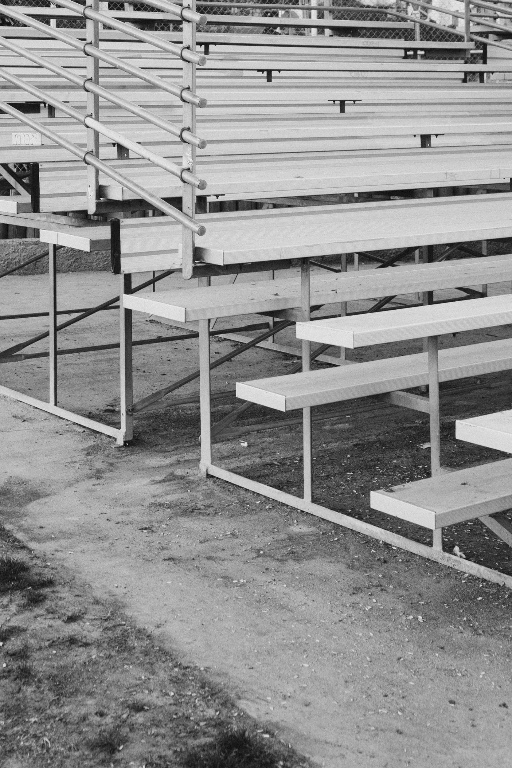 Empty bleacher seats on a dirt ground, metal railings, and a chain-link fence in the background.