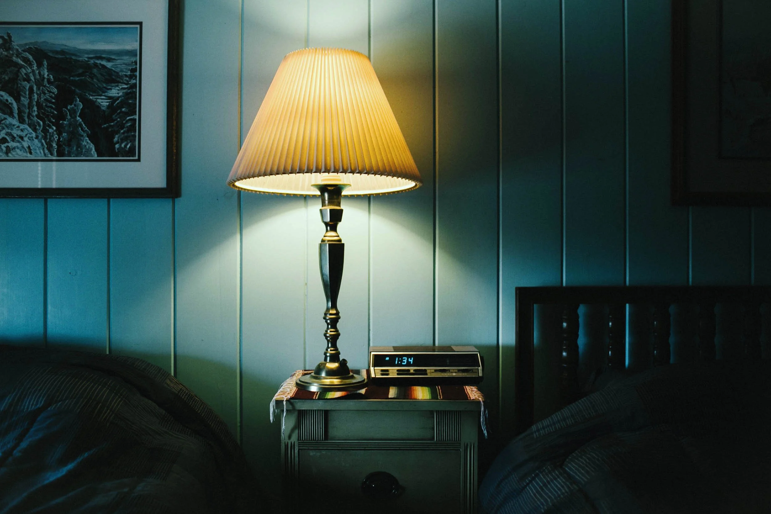 A bedroom bedside table with a lit lamp and digital clock showing 1:34, against paneled walls with framed artwork.