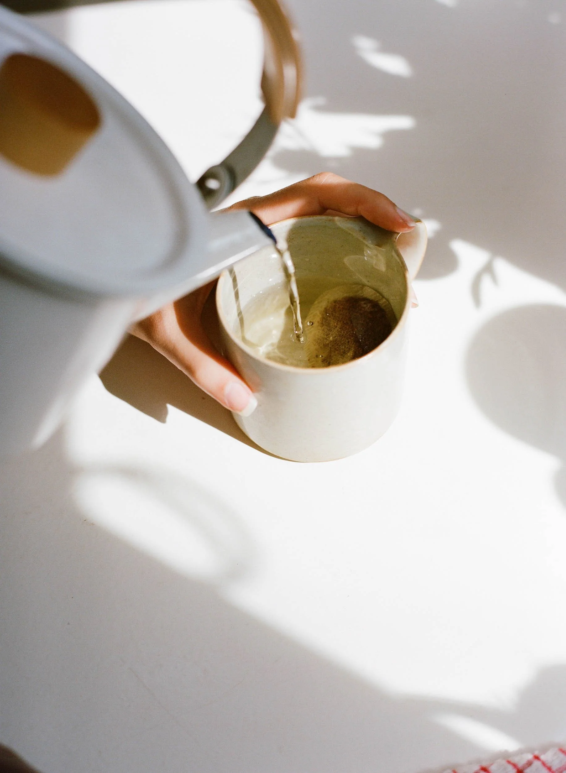 Pouring hot water into a ceramic mug placed on a white surface, with shadows and natural light creating a bright atmosphere.