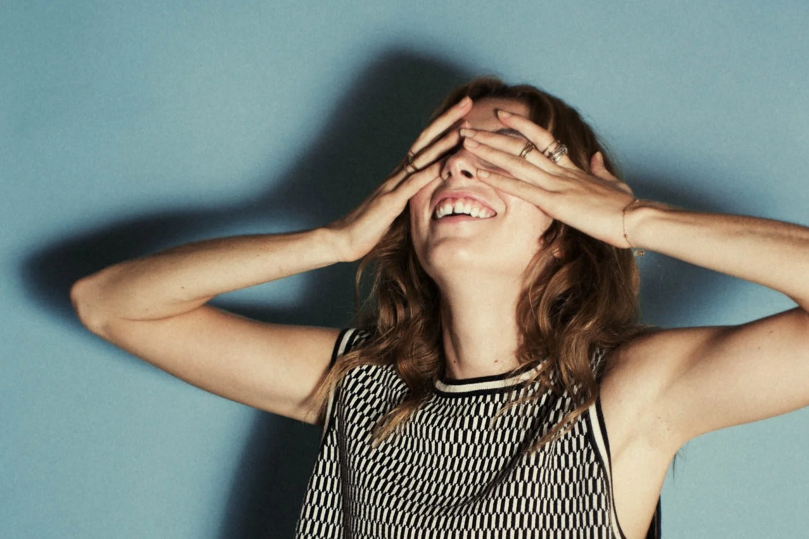 Woman smiling with her hands covering her eyes against a blue background.
