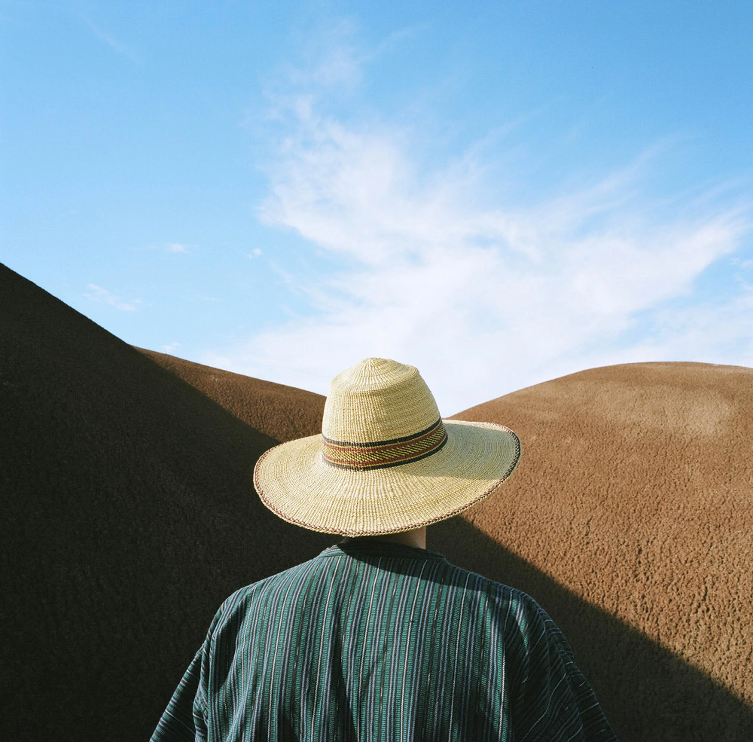 Back of a person wearing a wide-brimmed straw hat and striped shirt, standing between brown sand dunes with a blue sky and scattered clouds above.