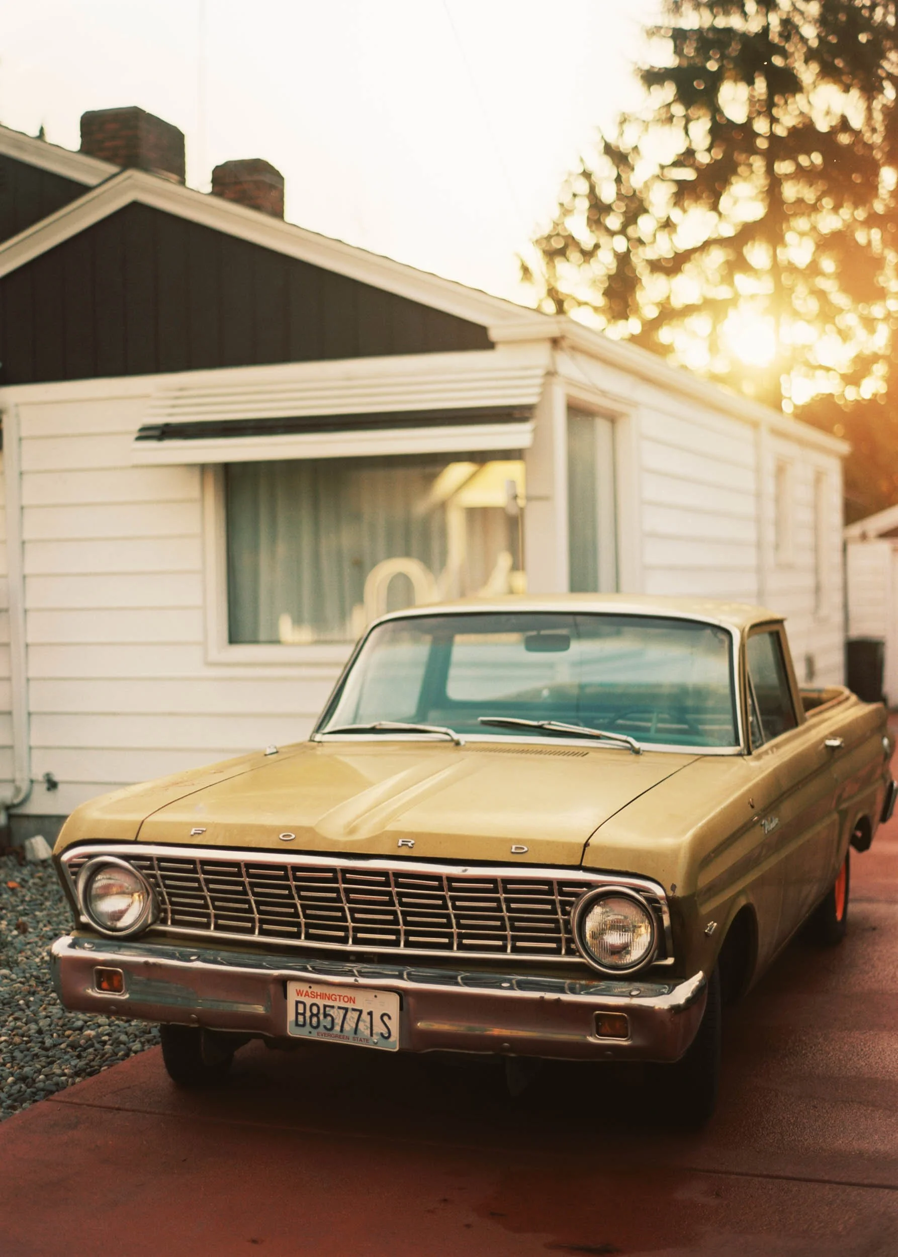 A vintage yellow Ford car parked in front of a white house with a large window, during sunset.