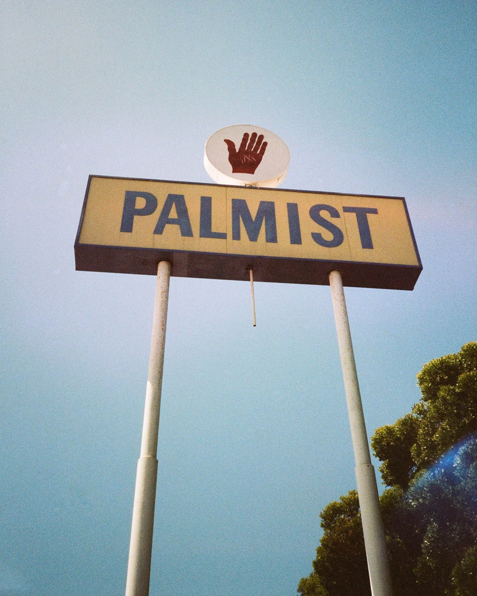 A sign with a yellow background and blue text reading 'PALM IST' is below a circular sign with a red hand symbol. The sign is mounted on white poles against a blue sky with some green trees visible at the bottom right.
