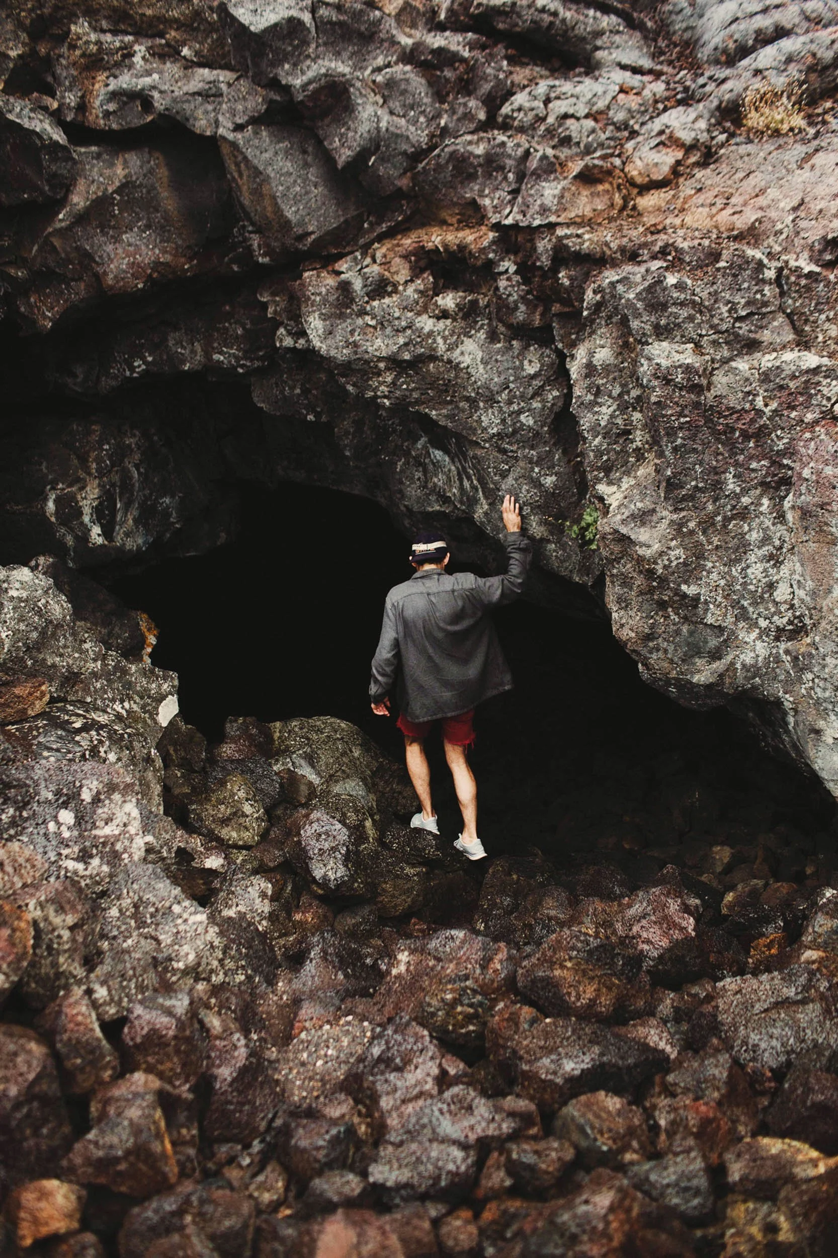 A man in a gray jacket, red shorts, and sneakers walking into a dark cave surrounded by large rocks.