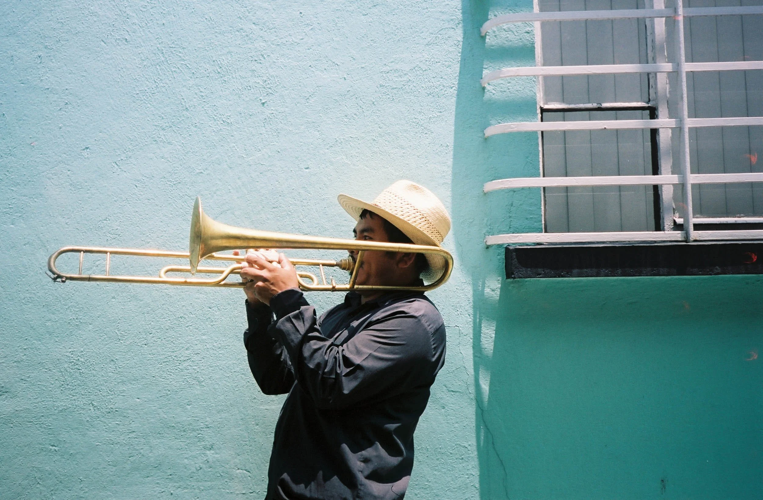 A person wearing a straw hat playing a trombone against a turquoise wall with a window and white security bars.