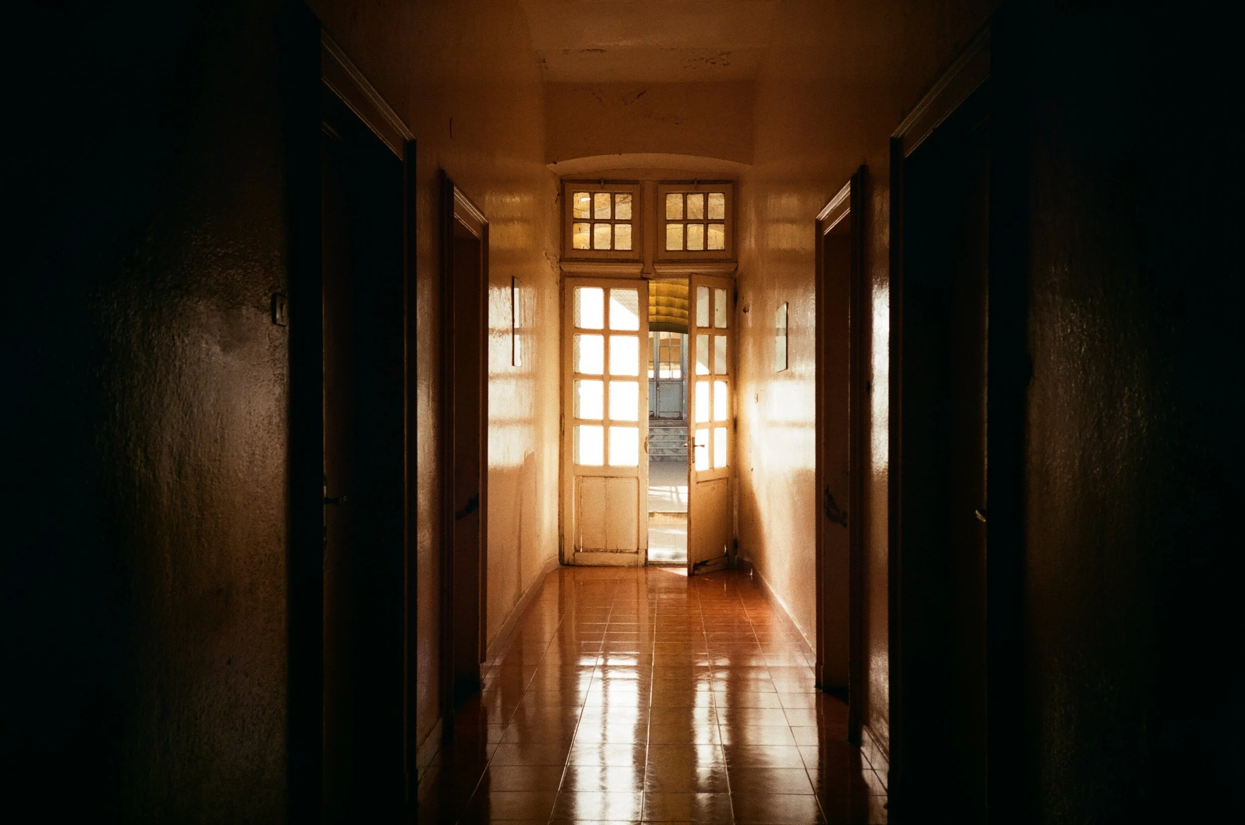 Dimly lit hallway with open glass double doors at the end, sunlight streaming through, tiled floor reflecting light.