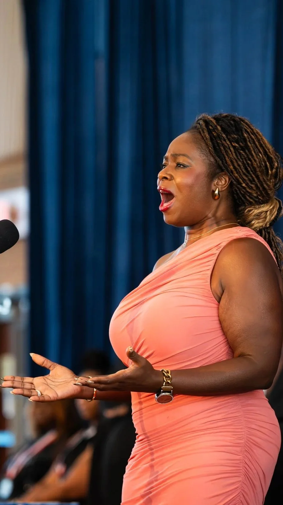 A woman with long braids, dressed in a pink sleeveless dress, speaking passionately with her hands outstretched in front of a blue curtain.