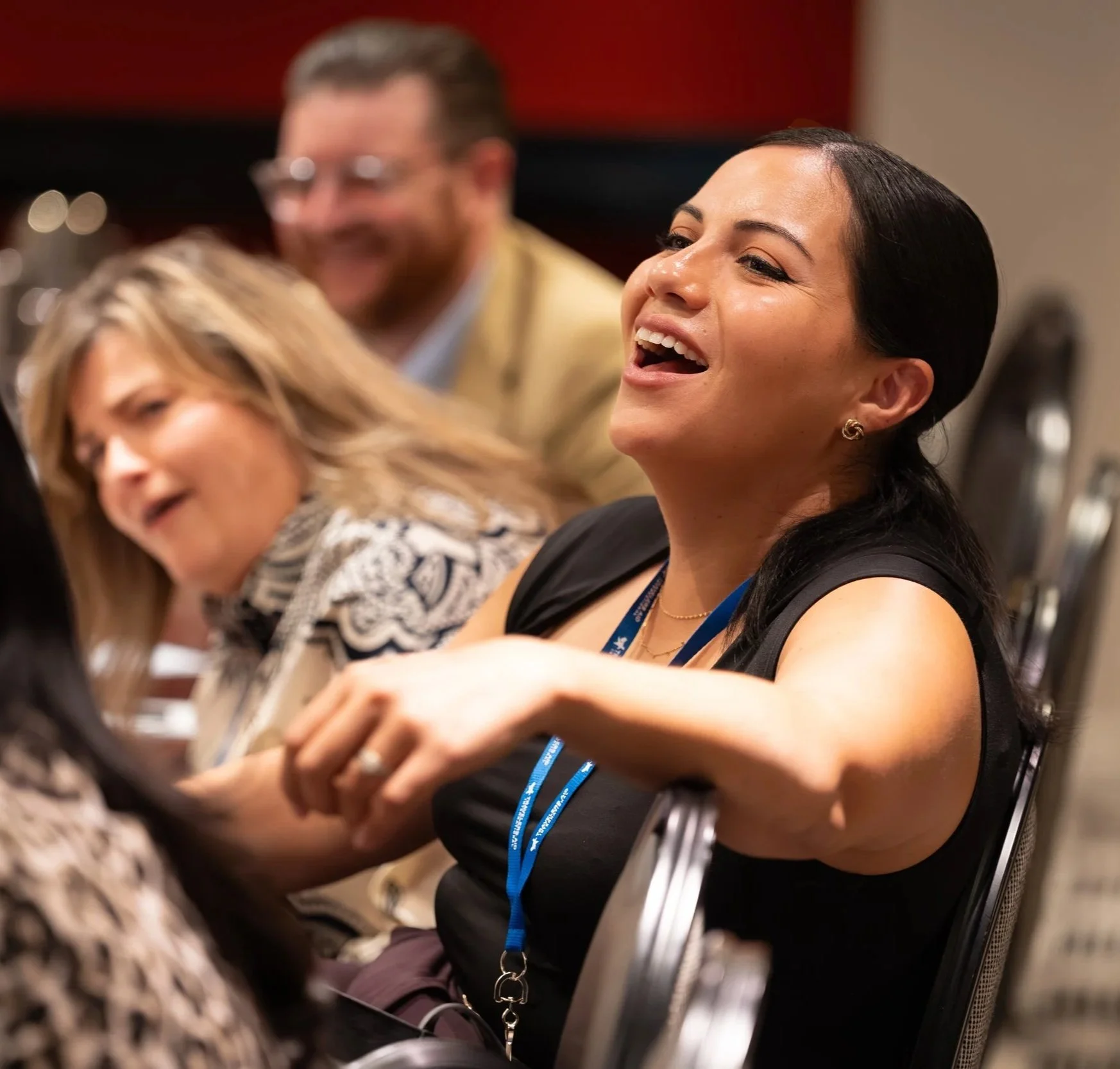 A woman with dark hair and earrings, wearing a black sleeveless top, happily laughing and holding hands with someone in a group at a conference or meeting.