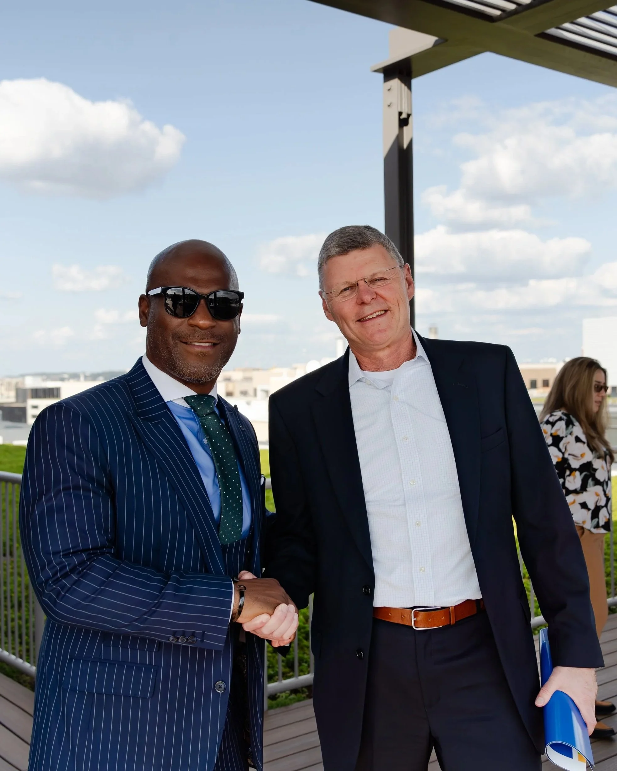 Two men in suits shaking hands outdoors on a rooftop, smiling, with a woman in sunglasses in the background, against a cityscape with buildings and a partly cloudy sky.