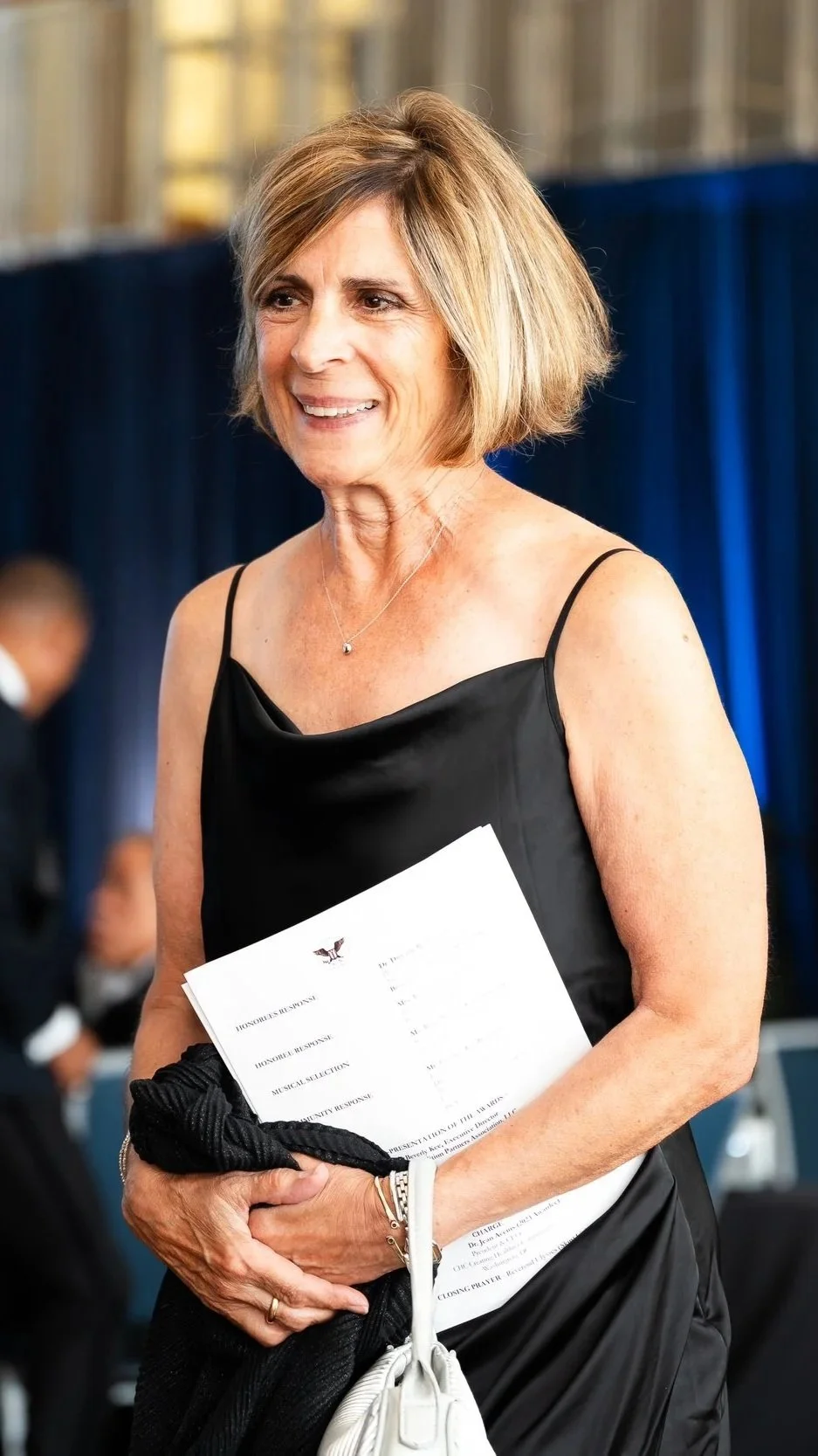A middle-aged woman with shoulder-length blonde hair wearing a black sleeveless dress, smiling, holding a paper program and a white bag at an event.