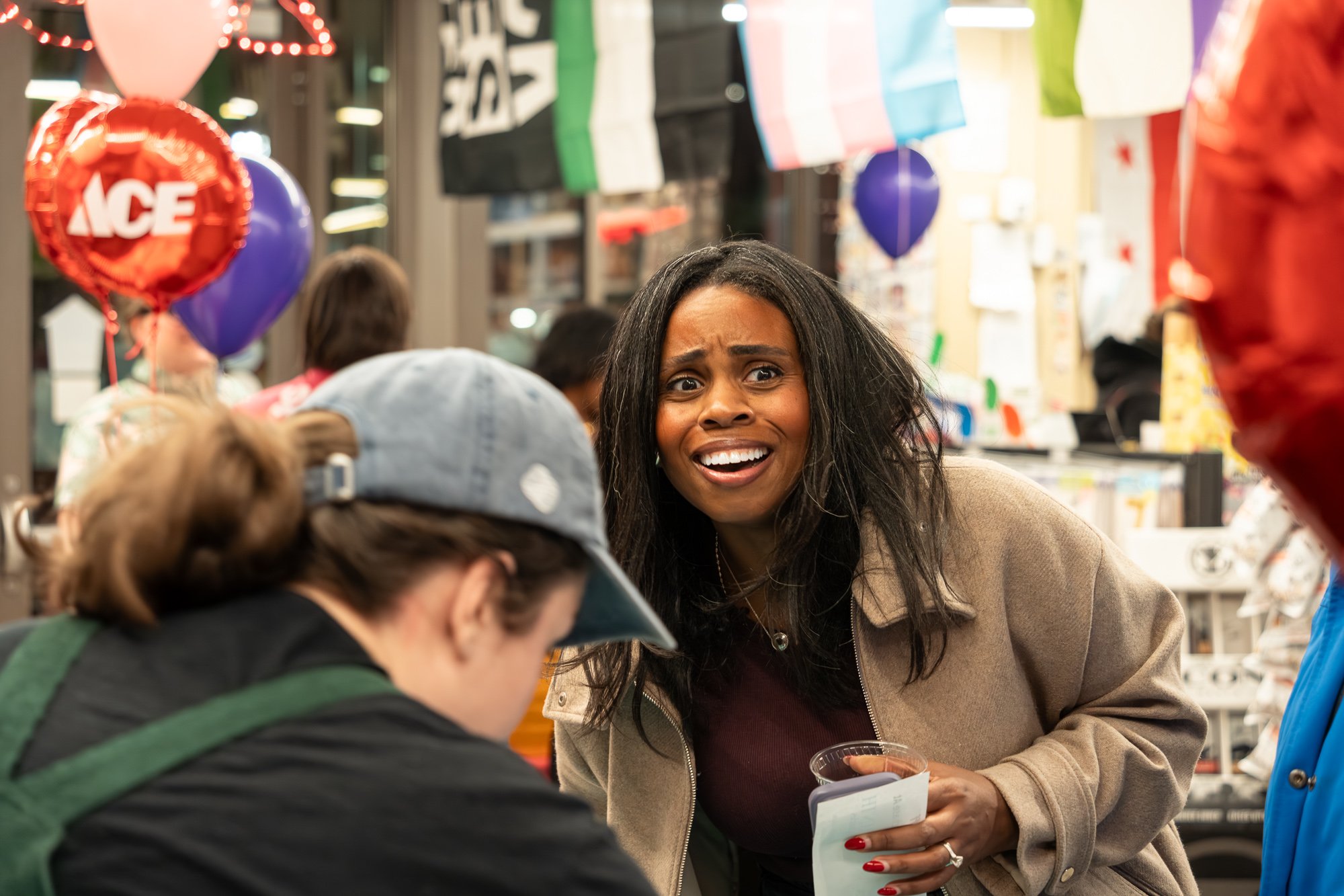 A woman looks surprised and confused while talking to a person at a checkout counter in a store decorated with balloons and flags.