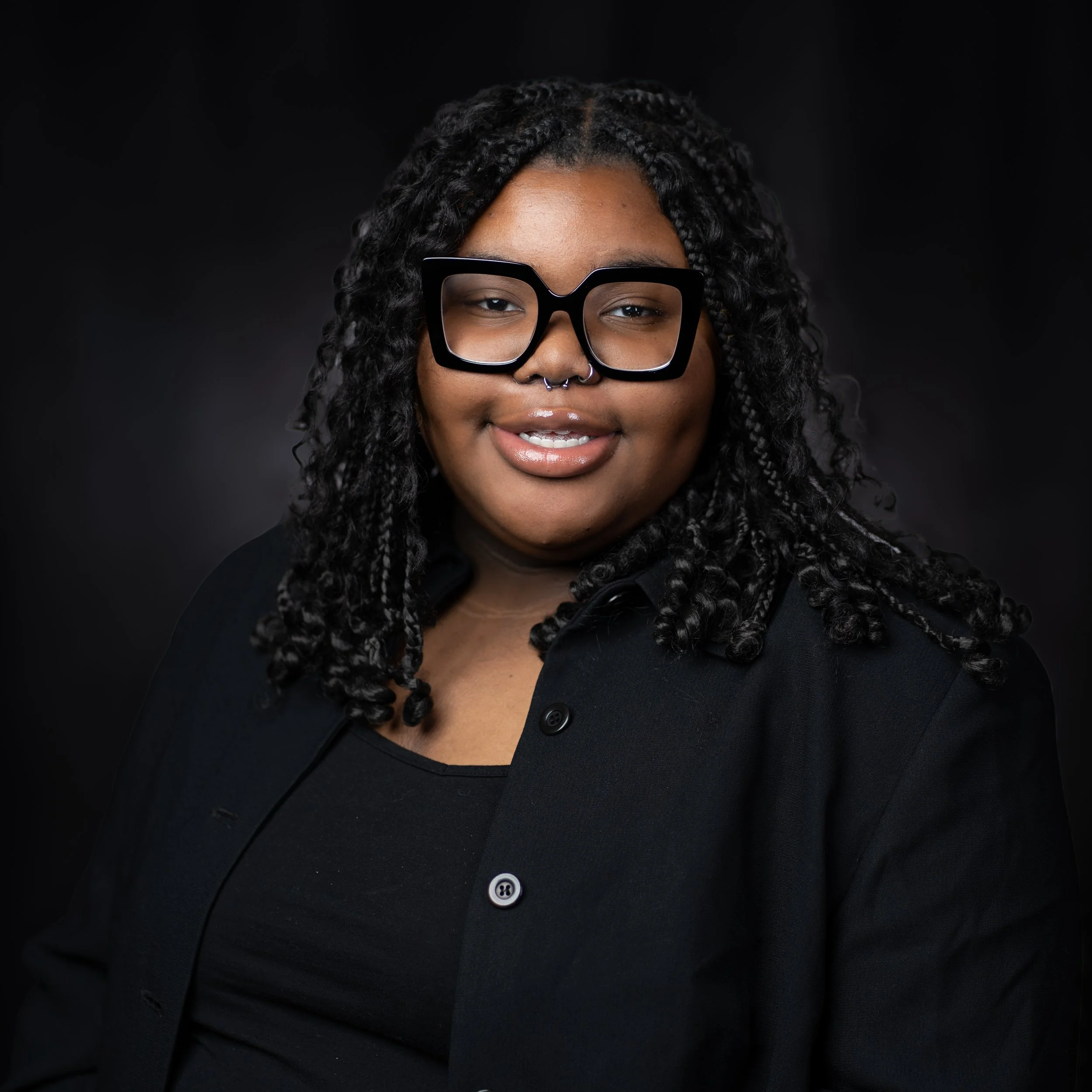 Portrait of a woman with dark curly hair, wearing large black glasses and a septum piercing, smiling against a dark background.