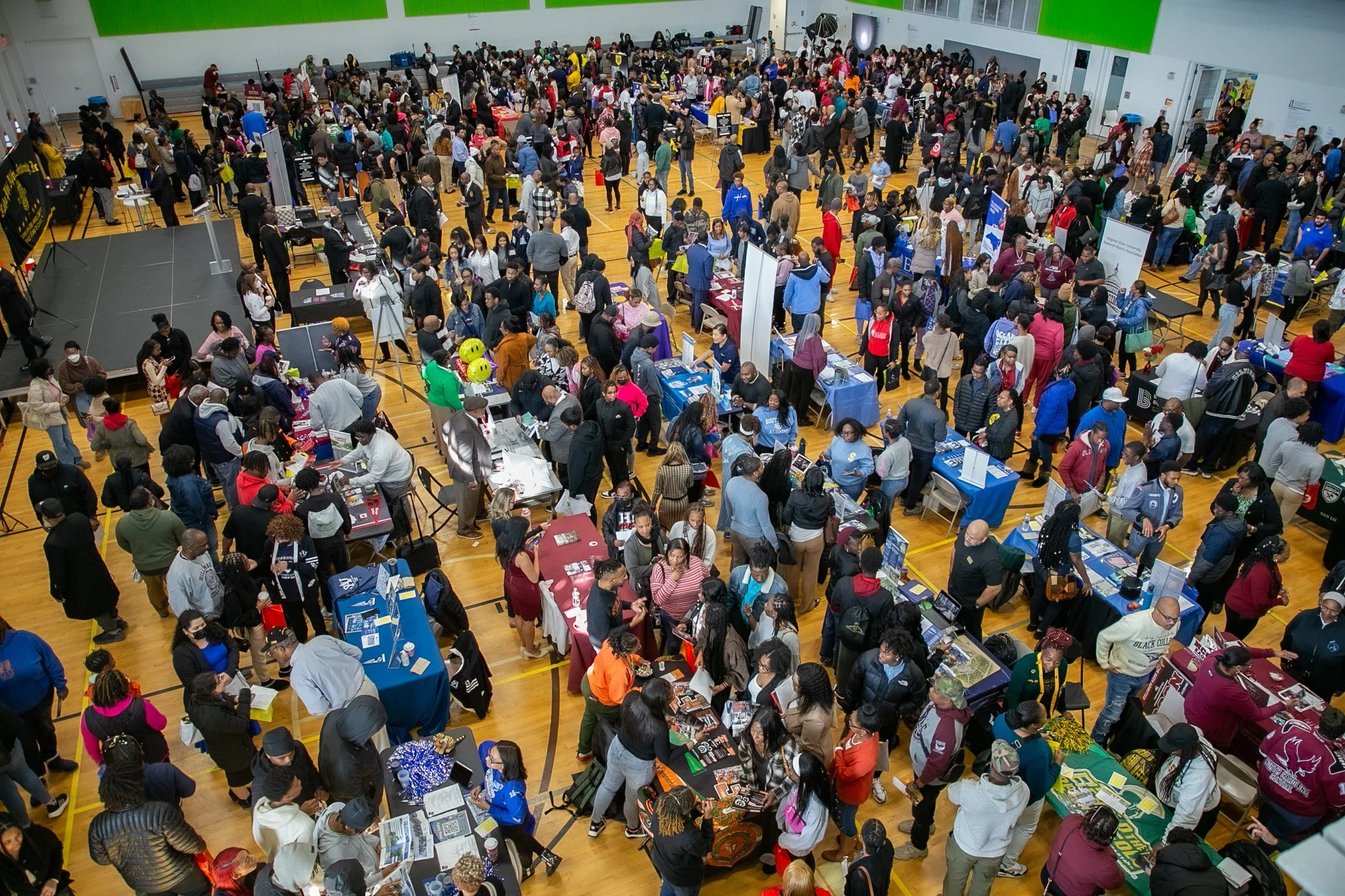 A crowded indoor event with numerous booths and tables, filled with people browsing and interacting in a gymnasium with high ceilings and wooden floors.