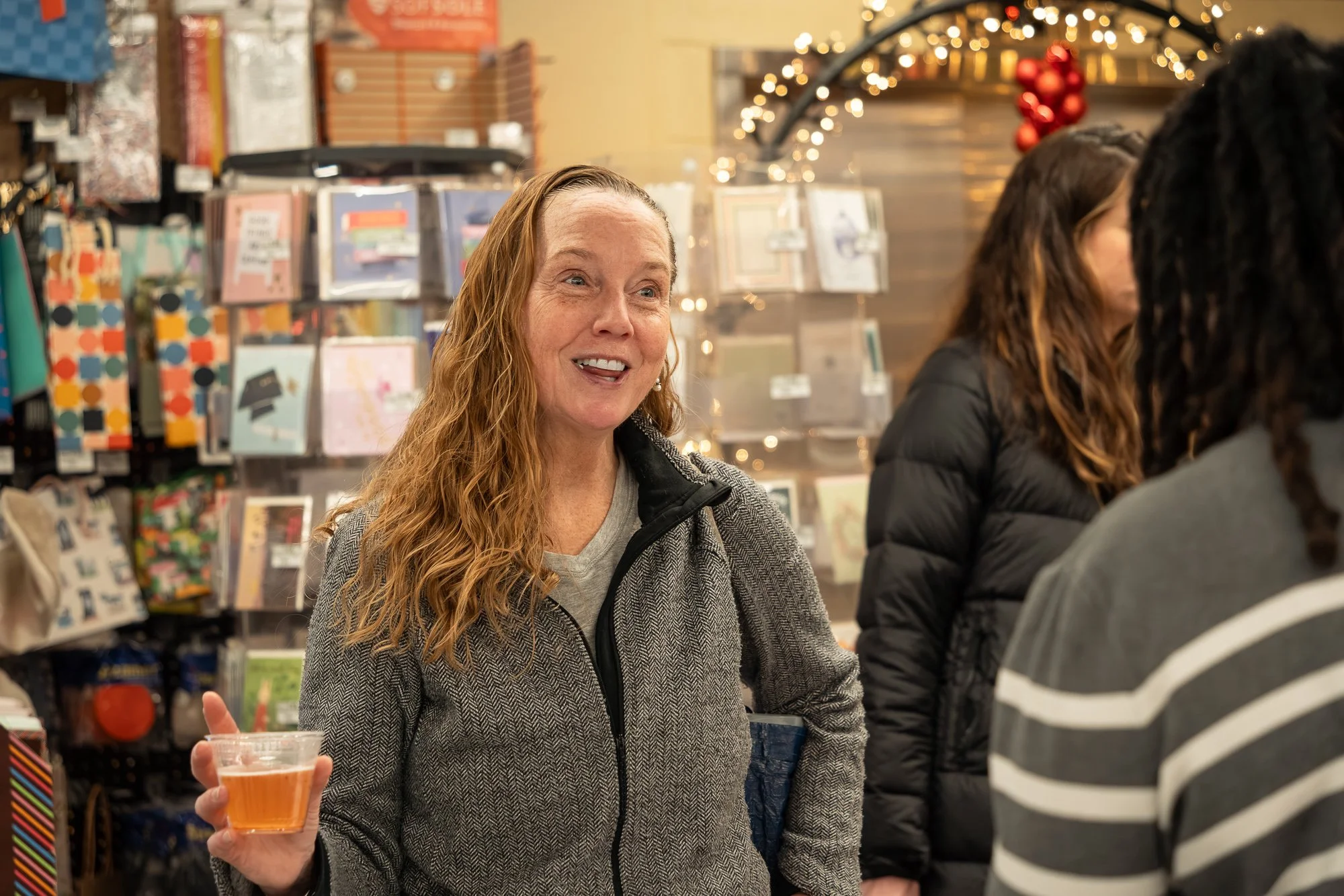 A woman with long red hair smiling and holding a drink in a store decorated for Christmas with holiday lights and ornaments.