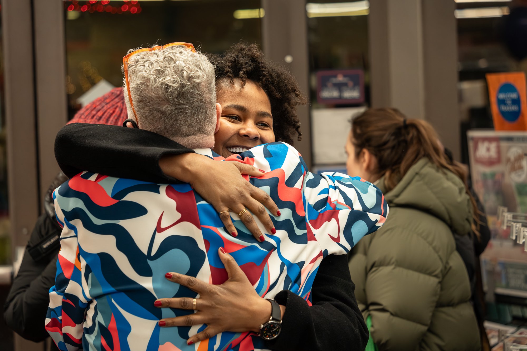 Two women hugging each other warmly in an indoor setting, with one smiling brightly, surrounded by people.