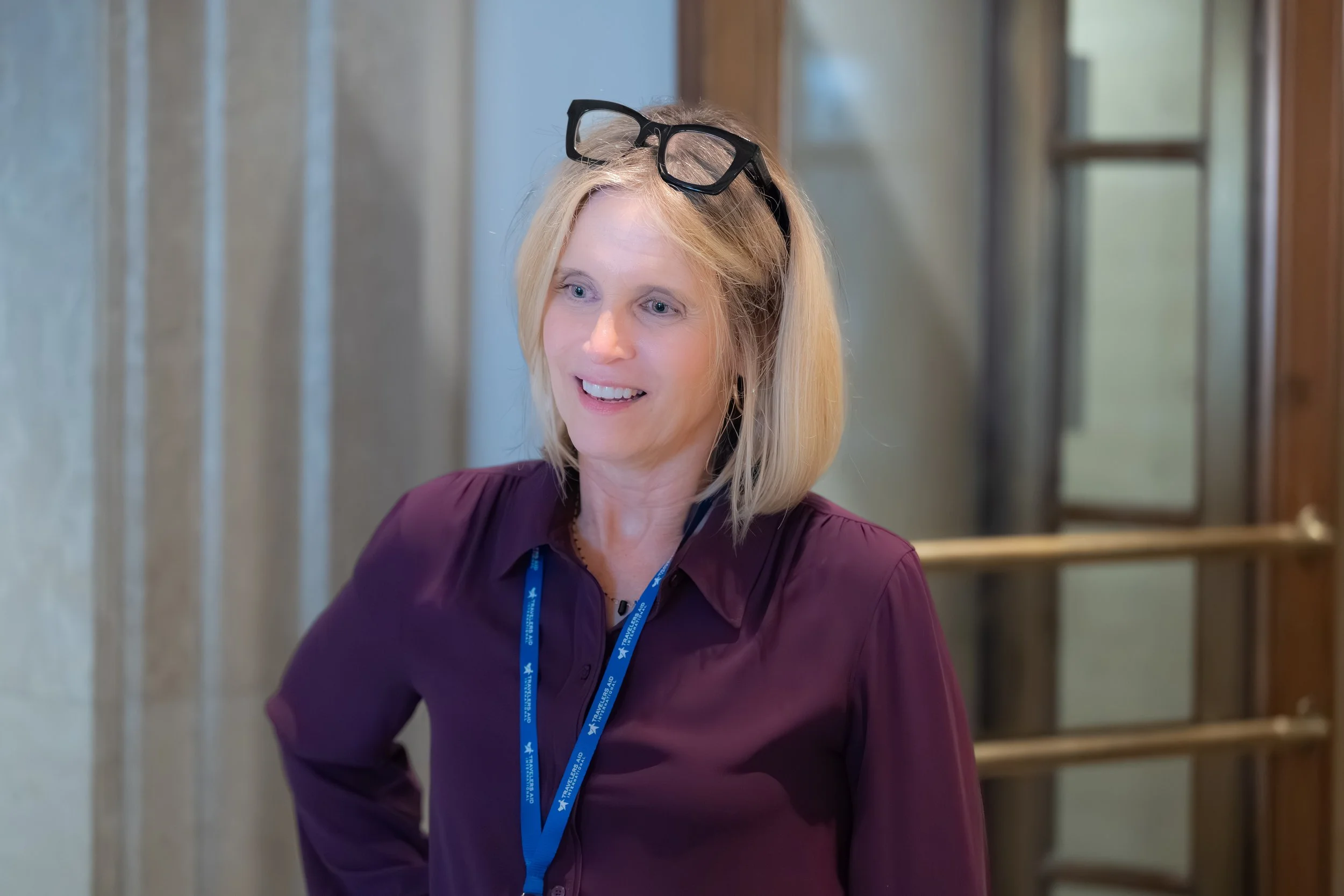 A blonde woman with glasses on her head, smiling and wearing a maroon blouse, standing indoors with wooden and glass doors in the background.