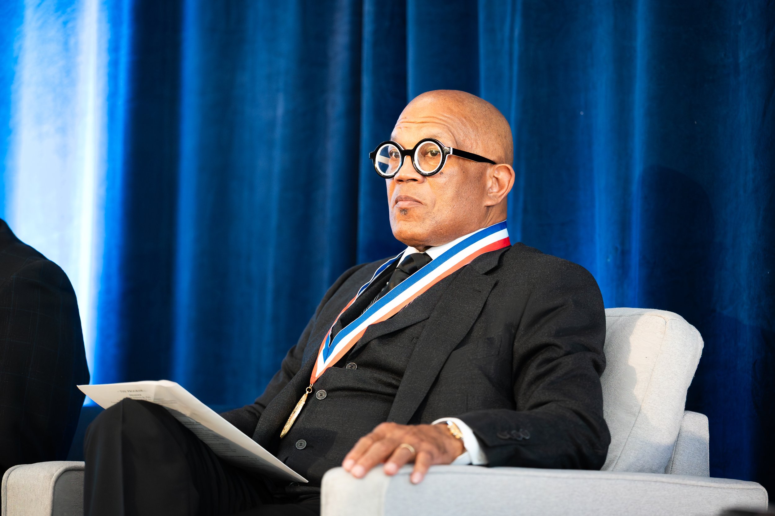 A bald man wearing glasses, a black suit, holding a paper, and wearing a medal with red, white, and blue stripes, sitting on a light-colored chair in front of a dark blue curtain.