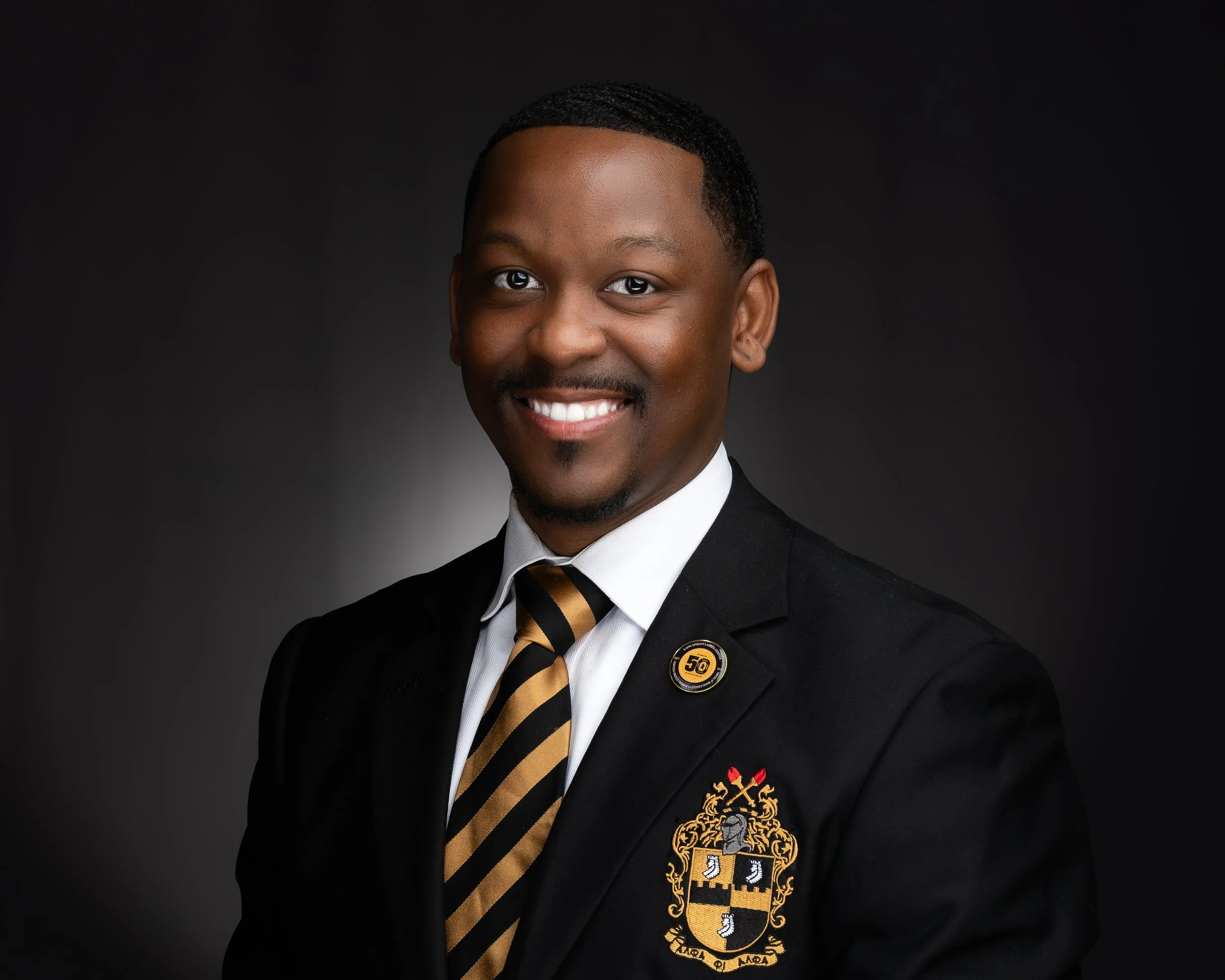 Portrait of a smiling man in a formal black suit with a crest on the chest, wearing a white shirt and a black and gold striped tie, against a dark background.