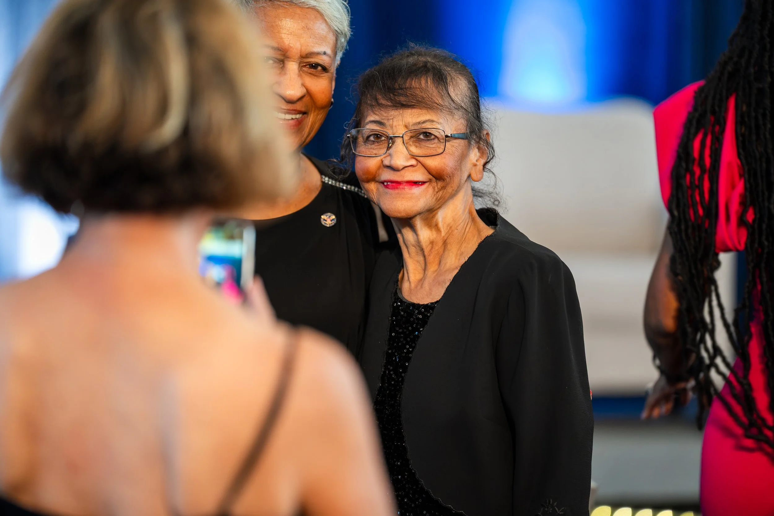 Three women are taking a photo together at an event. The woman in the center, elderly with glasses, is smiling and dressed in black. The woman on the left is partially visible and is wearing a black dress. The woman on the right has long black braids