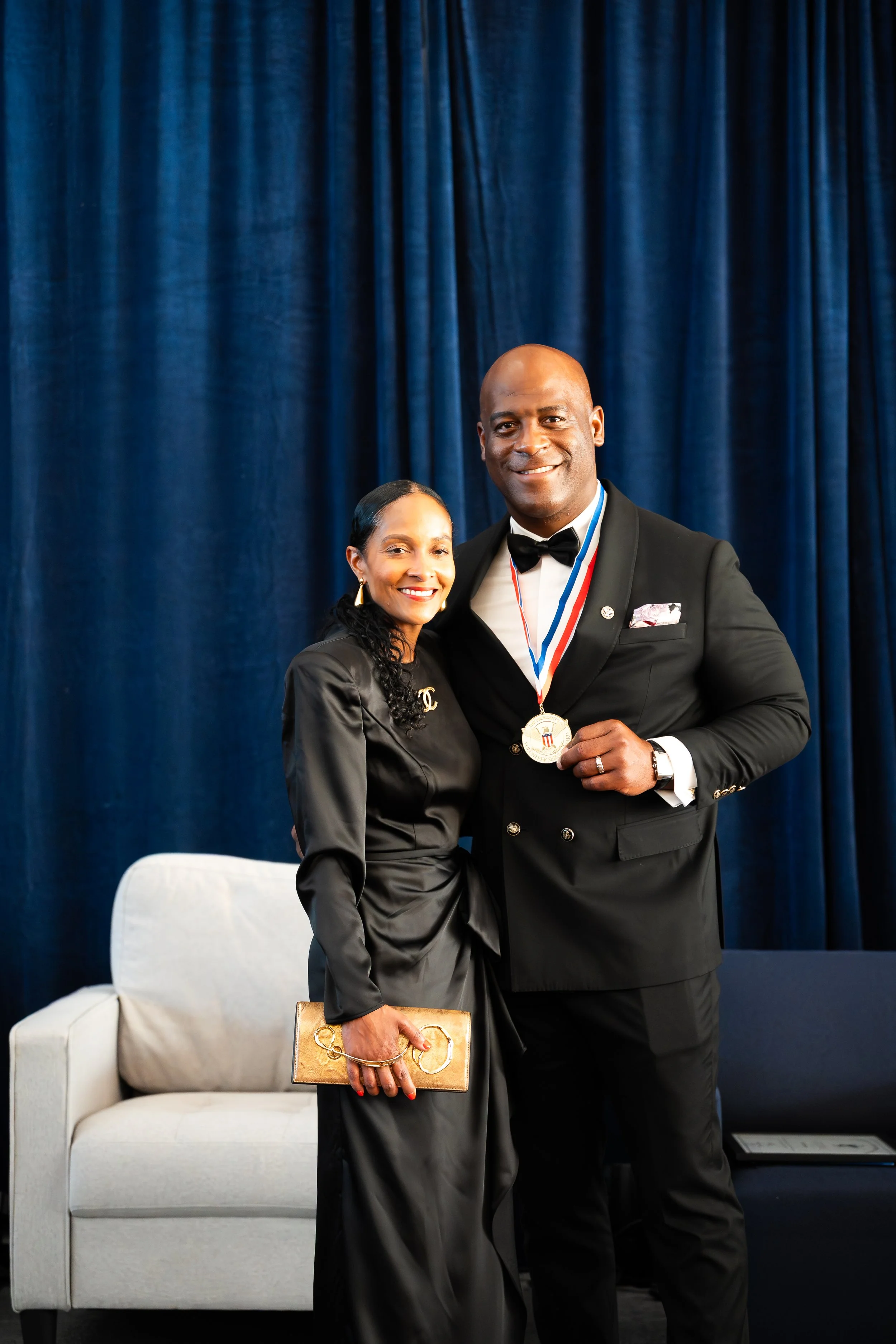 A man and woman dressed in formal attire smiling on stage, with the man wearing a medal around his neck, blue curtain backdrop, and a beige sofa behind them.