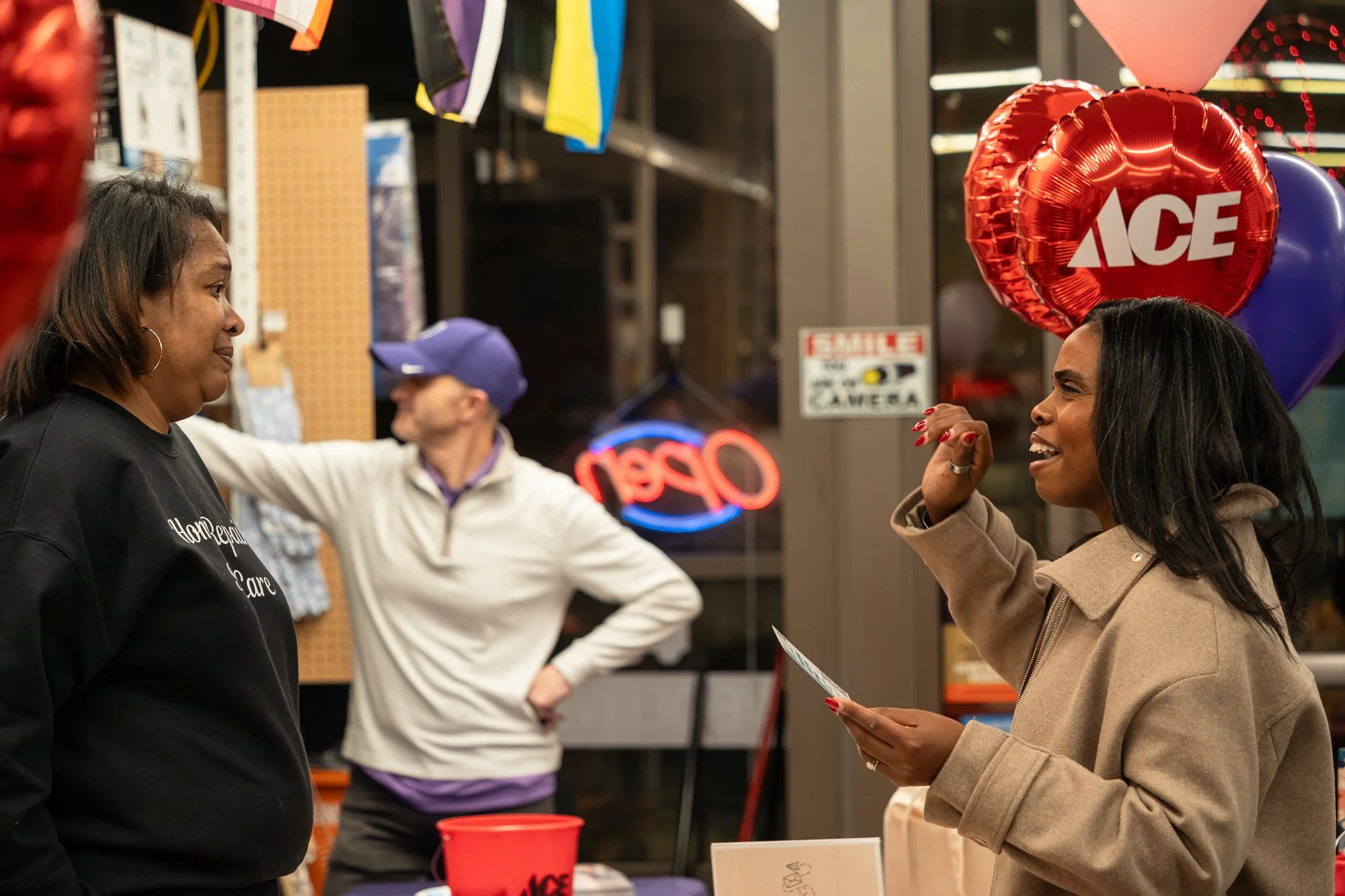 Two women talking at an indoor store or market, with balloons and neon signs in the background, one woman is holding a slip of paper.