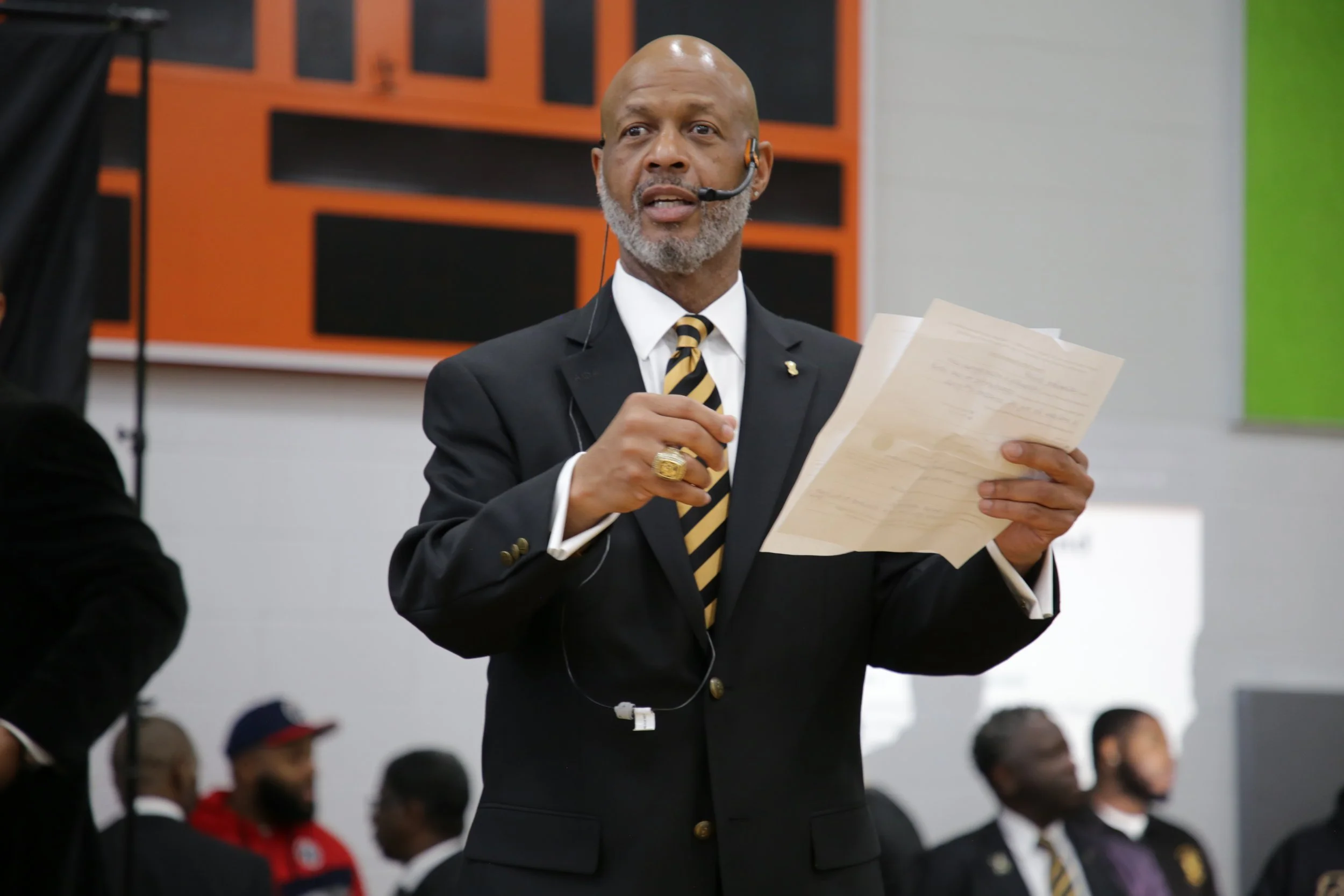 A man in a suit, with a gold ring and a striped tie, holding papers and speaking into a headset microphone at an indoor event.