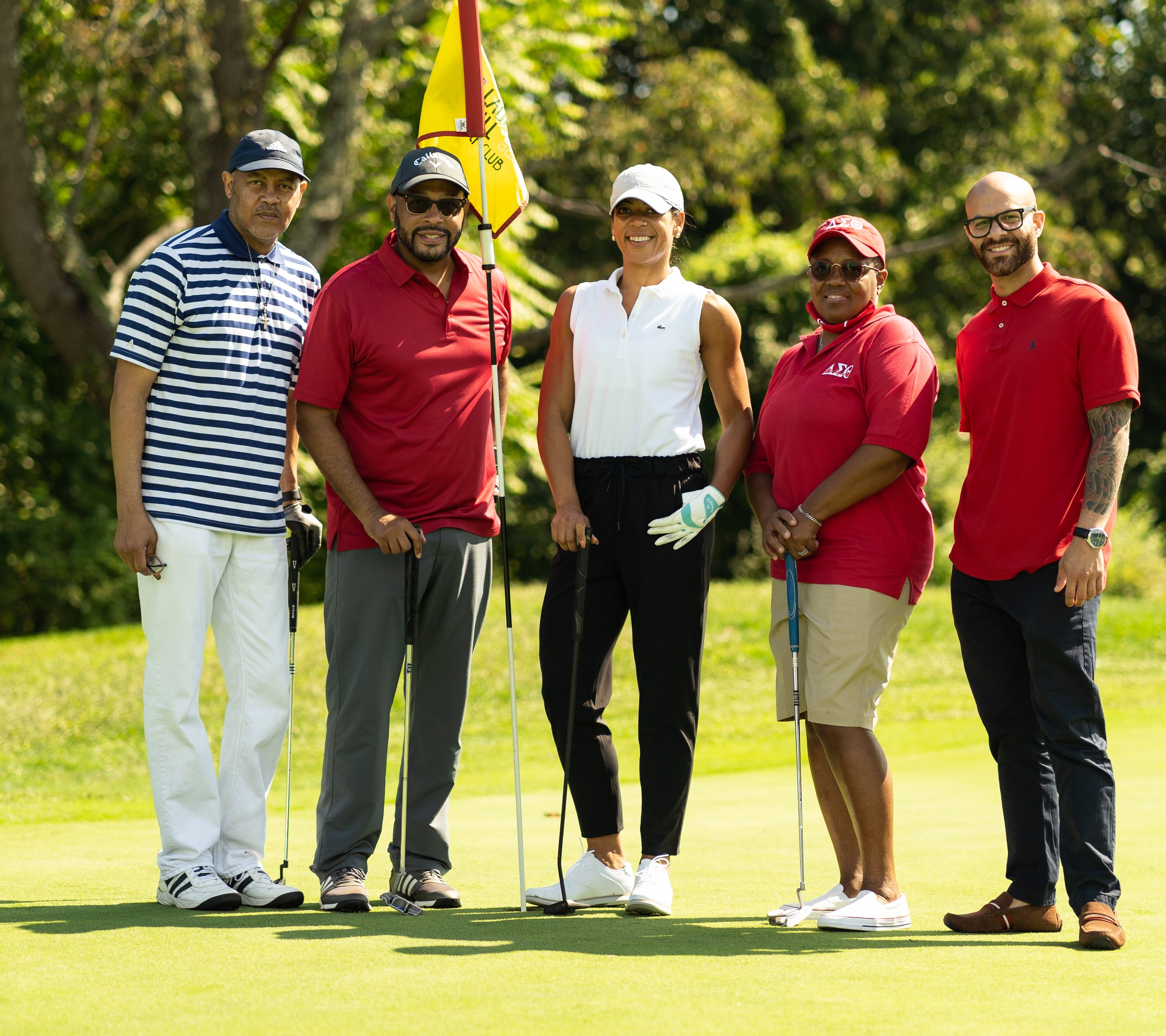 Group of five diverse people standing on a golf course, holding golf clubs, with trees in the background.