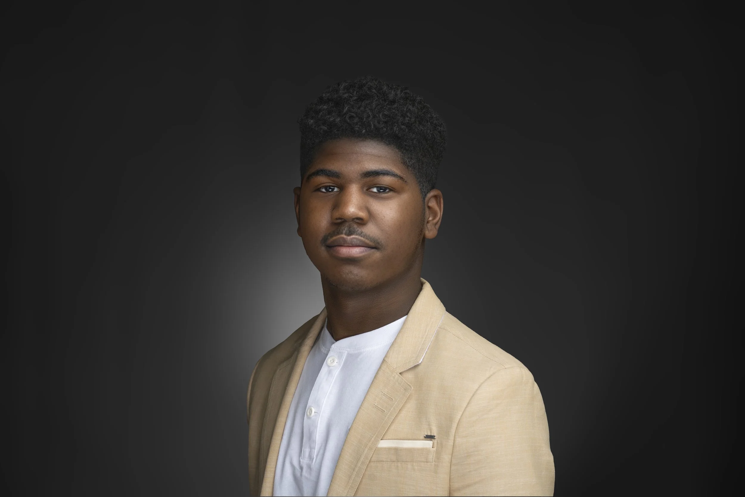 Portrait of a young man with dark curly hair, wearing a beige blazer over a white shirt, against a dark background.