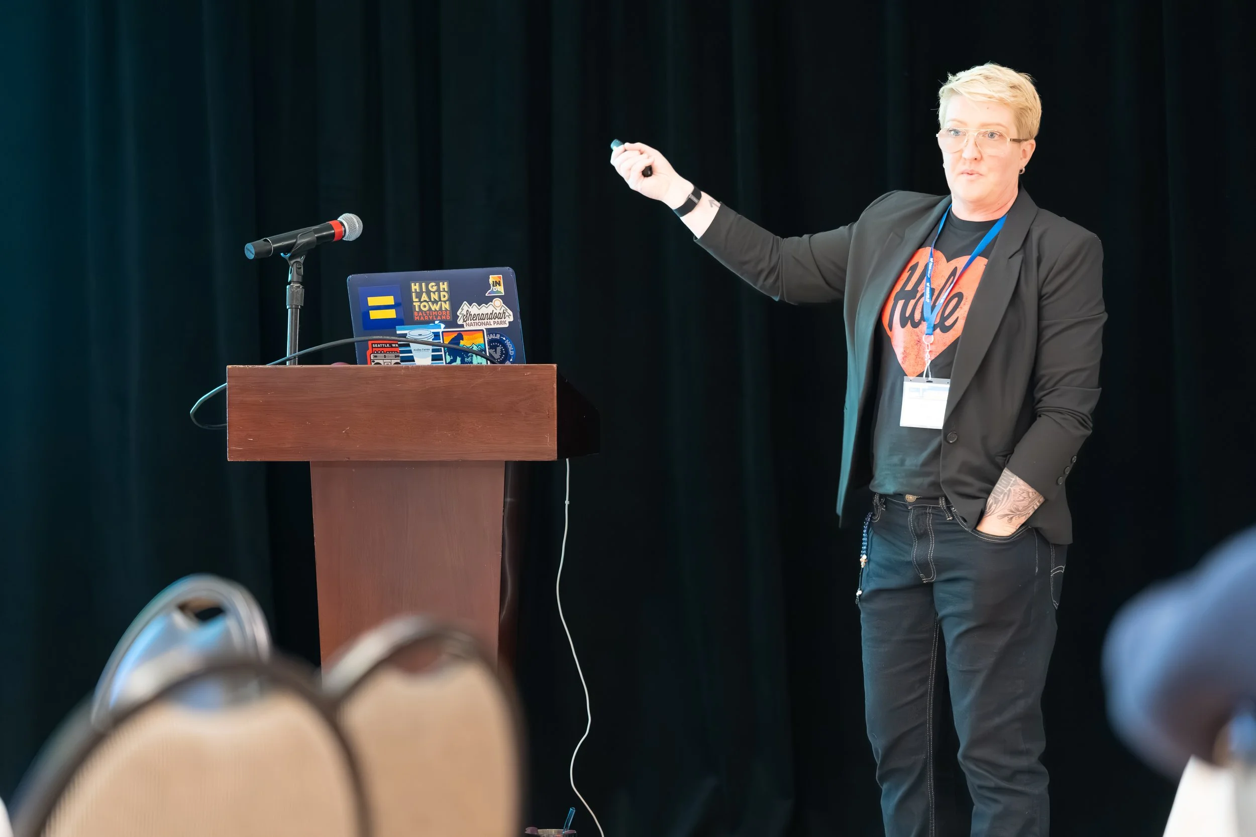 Person giving a presentation at a conference, standing next to a podium with a laptop decorated with stickers, wearing a blazer over a graphic T-shirt, holding a remote, with a dark curtain background.