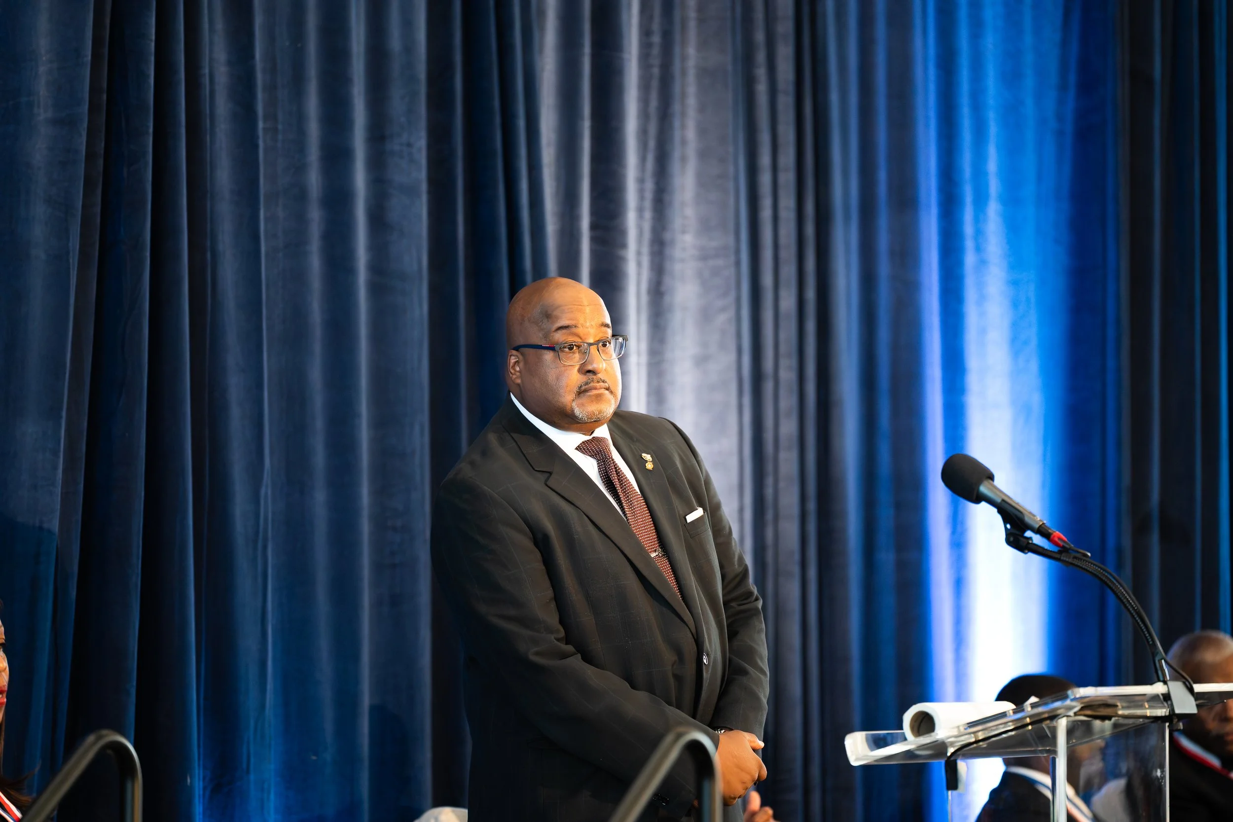 A man in a dark suit and glasses standing next to a podium with a microphone, against a backdrop of dark blue curtains.