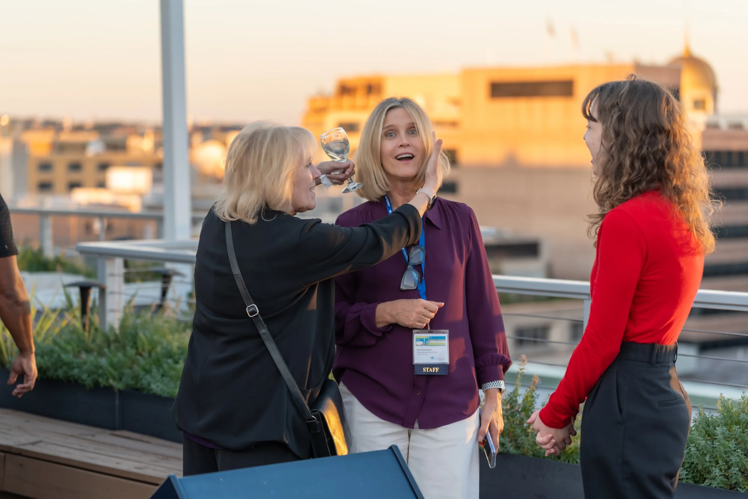 Three women are talking on a rooftop during sunset, one woman is playfully holding a glass of wine near another woman's face, while the third woman looks surprised.